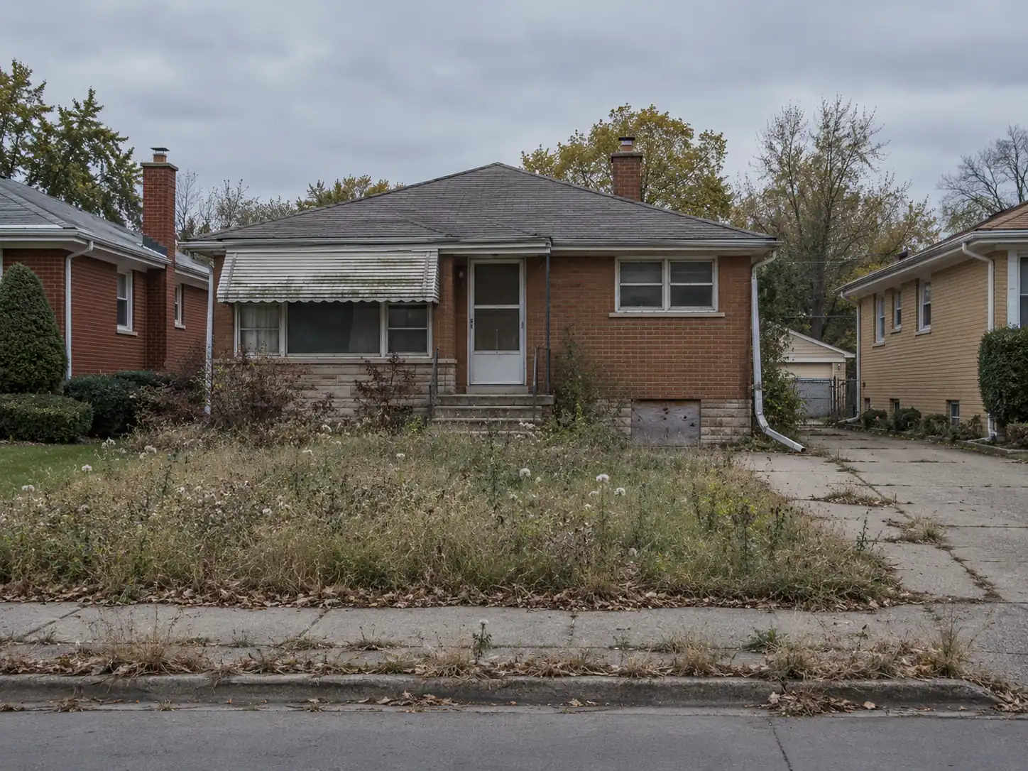Vacant brick ranch house with an overgrown front yard and a boarded basement window on an older Midwestern residential street