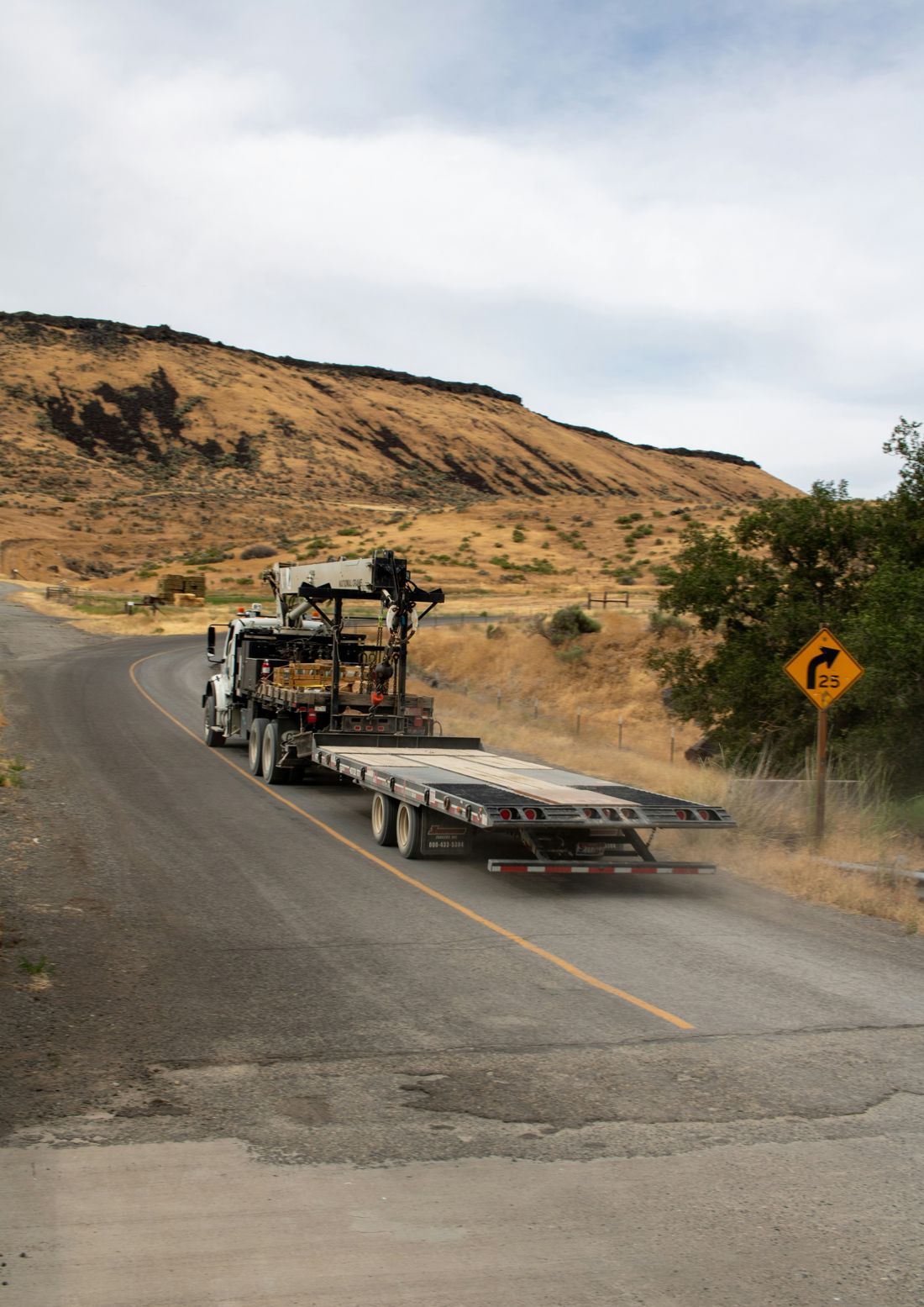 Flatbed truck hauling a crane down a winding rural road