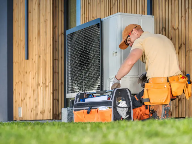 Technician servicing an outdoor heat pump unit with a tool bag.