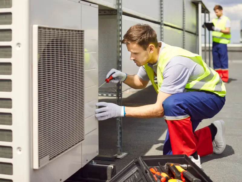 Two HVAC technicians inspecting a rooftop condenser unit with tools.