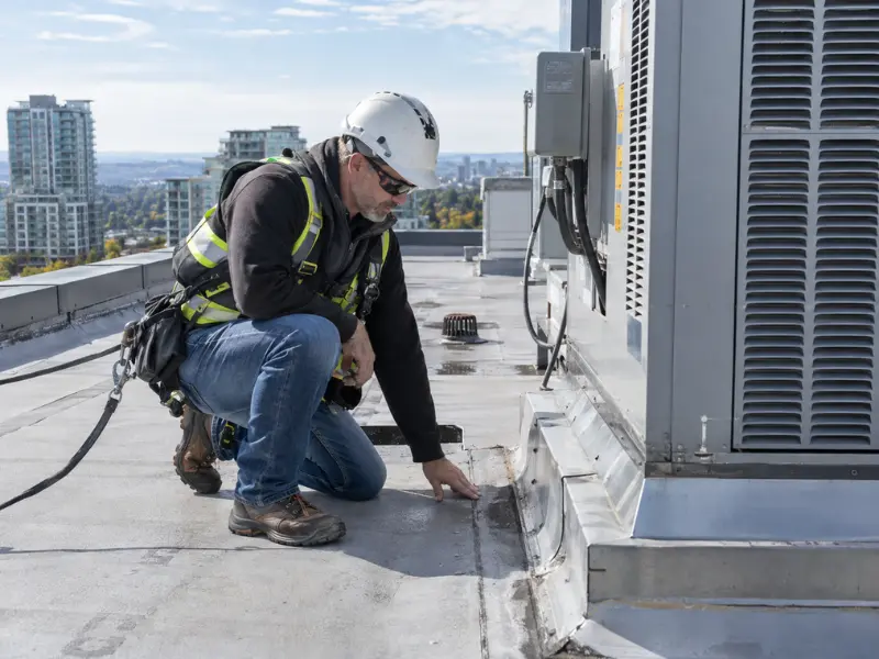 Building inspector in a hard hat and safety harness kneeling on a commercial rooftop to inspect flashing and an HVAC unit. Building inspector in a hard hat and safety harness kneeling on a commercial rooftop to inspect flashing and an HVAC unit.