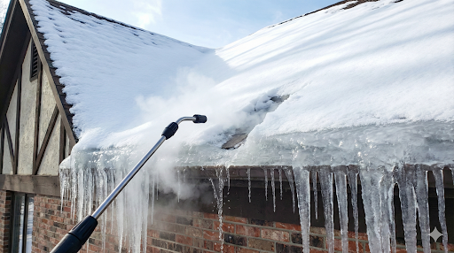 Professional steam equipment being used to remove a thick ice dam and icicles from a residential roof gutter.