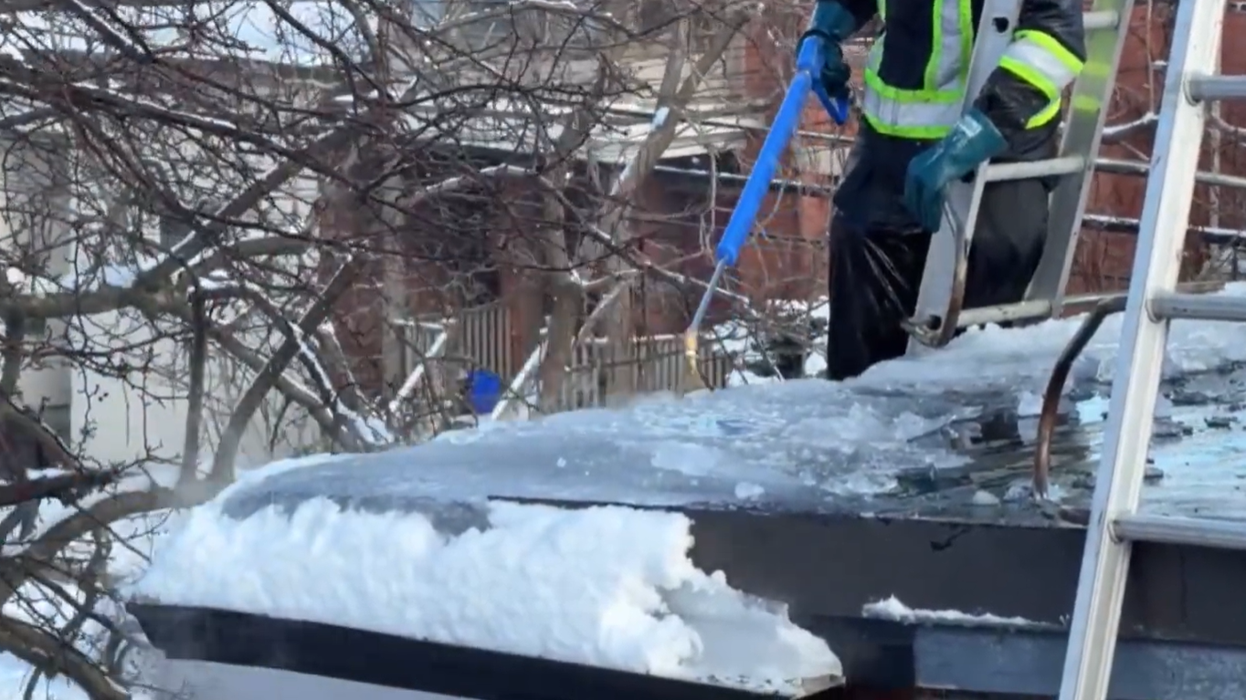 A worker wearing protective gear and high-visibility clothing uses a specialized tool to steam and melt ice off a snow-covered roof, with a metal ladder visible in the foreground.