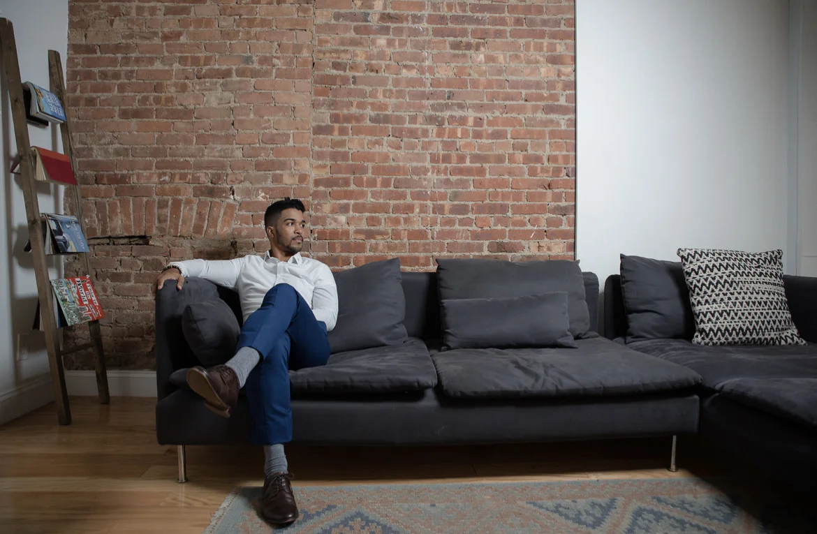 Ralph Llenas Ruiz sitting on a sofa in front of a brick wall, looking to one side