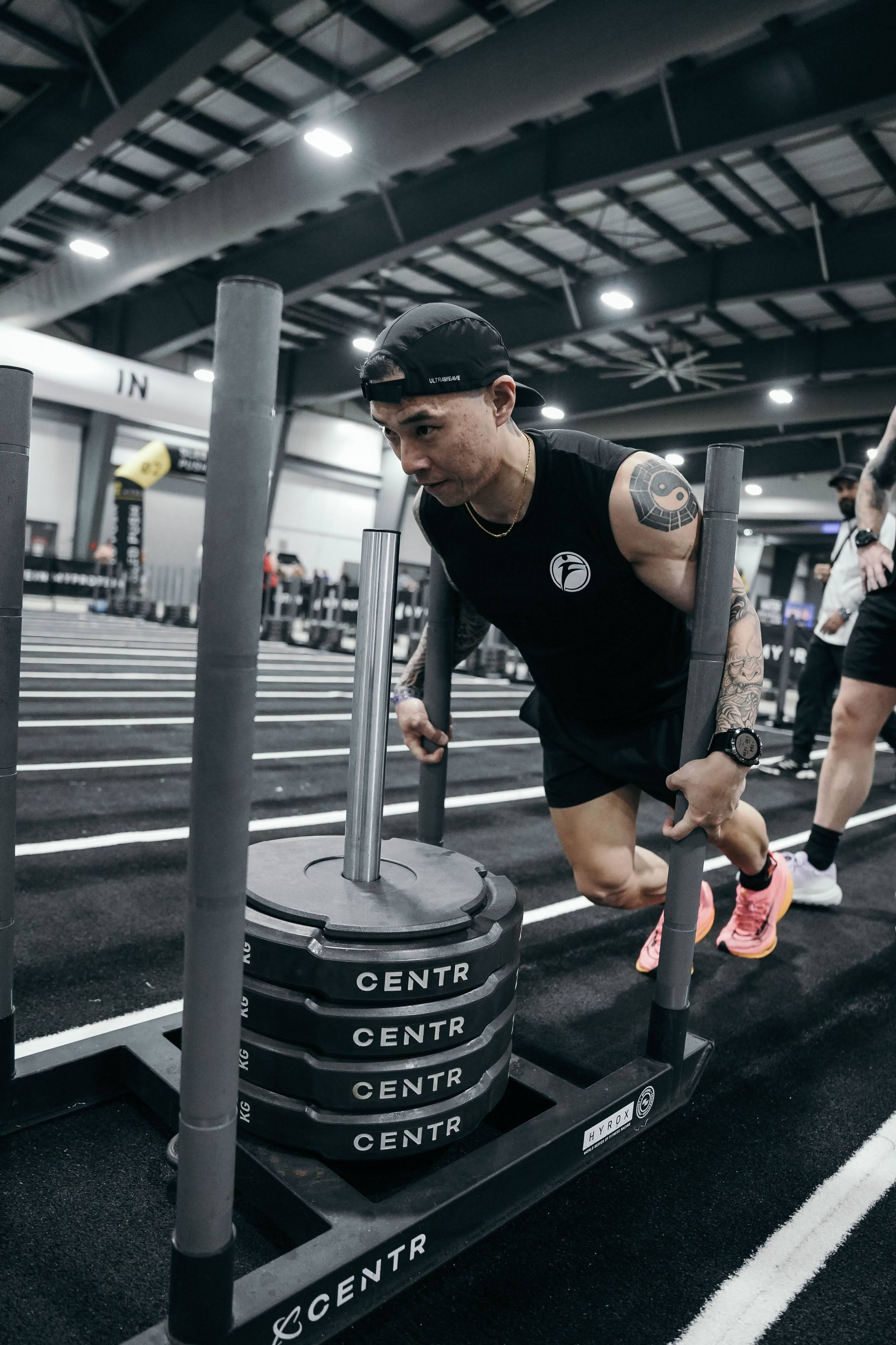 Joe Hsiung pushing a weighted sled on the carpeted track at the Hyrox Ottawa 2026 Open Doubles competition at the EY Centre. He is in a low driving position with both hands gripping the sled handles, visibly straining against the resistance of the competition surface.