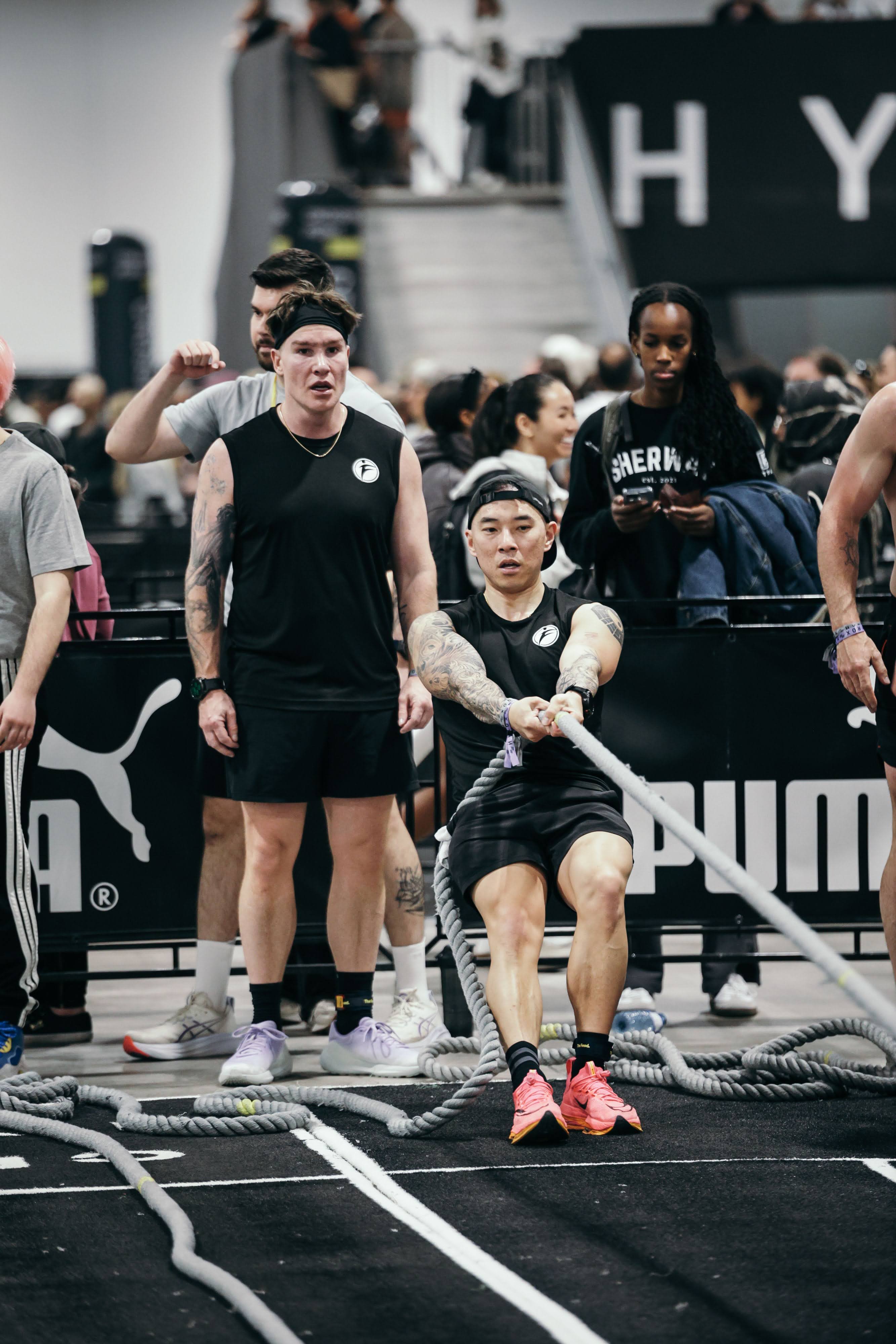 Joe Hsiung pulling a weighted sled using a rope at the Hyrox Ottawa 2026 Open Doubles competition. He is seated on the ground in the designated lane, pulling the rope hand over hand to drag the sled toward him inside the EY Centre arena.
