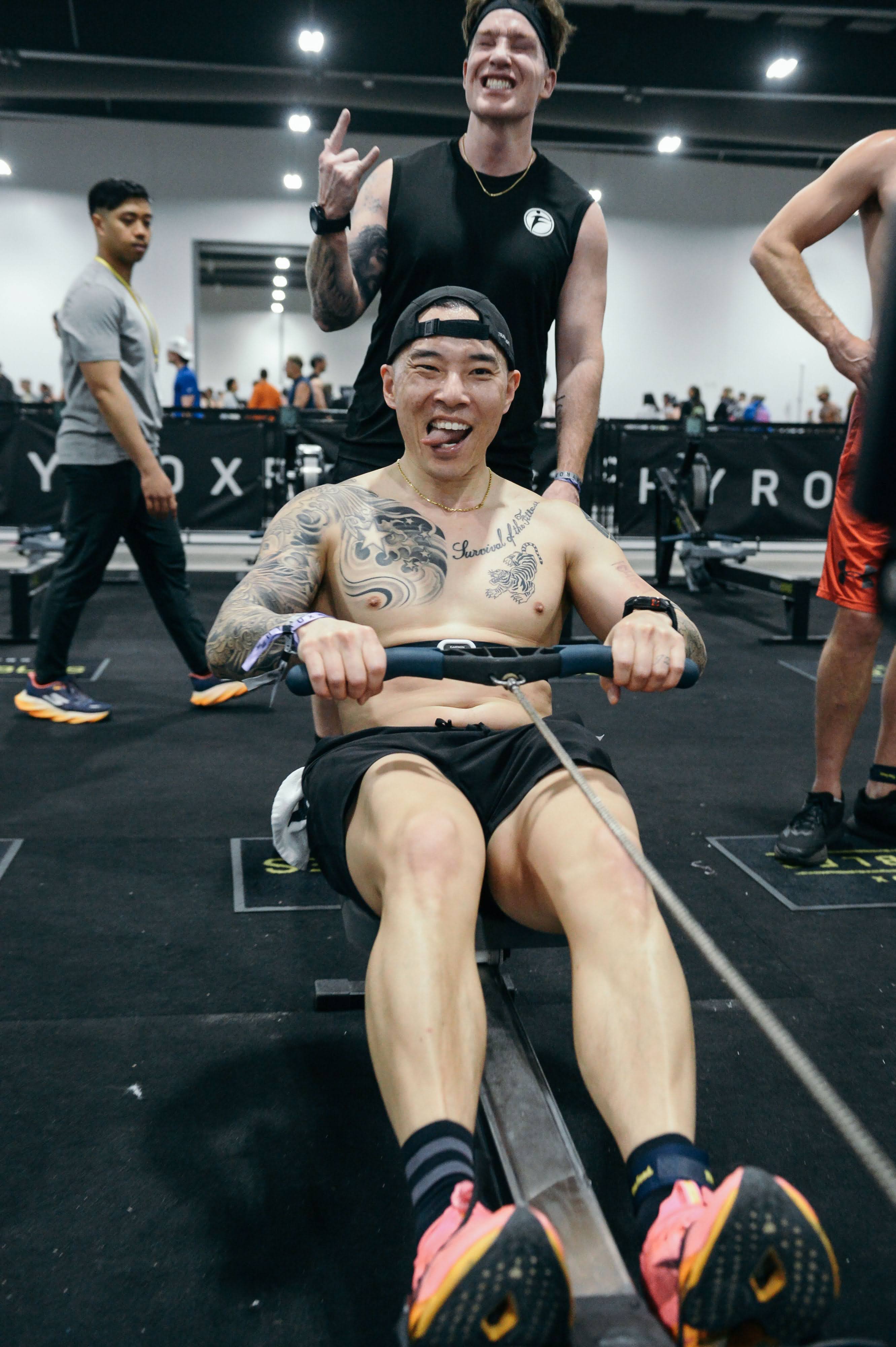 Joe Hsiung on the rowing machine at the Hyrox Ottawa 2026 Open Doubles competition. He is mid-stroke with an intense expression, pulling the handle toward his chest inside the EY Centre arena with other competitors and Hyrox signage in the background.
