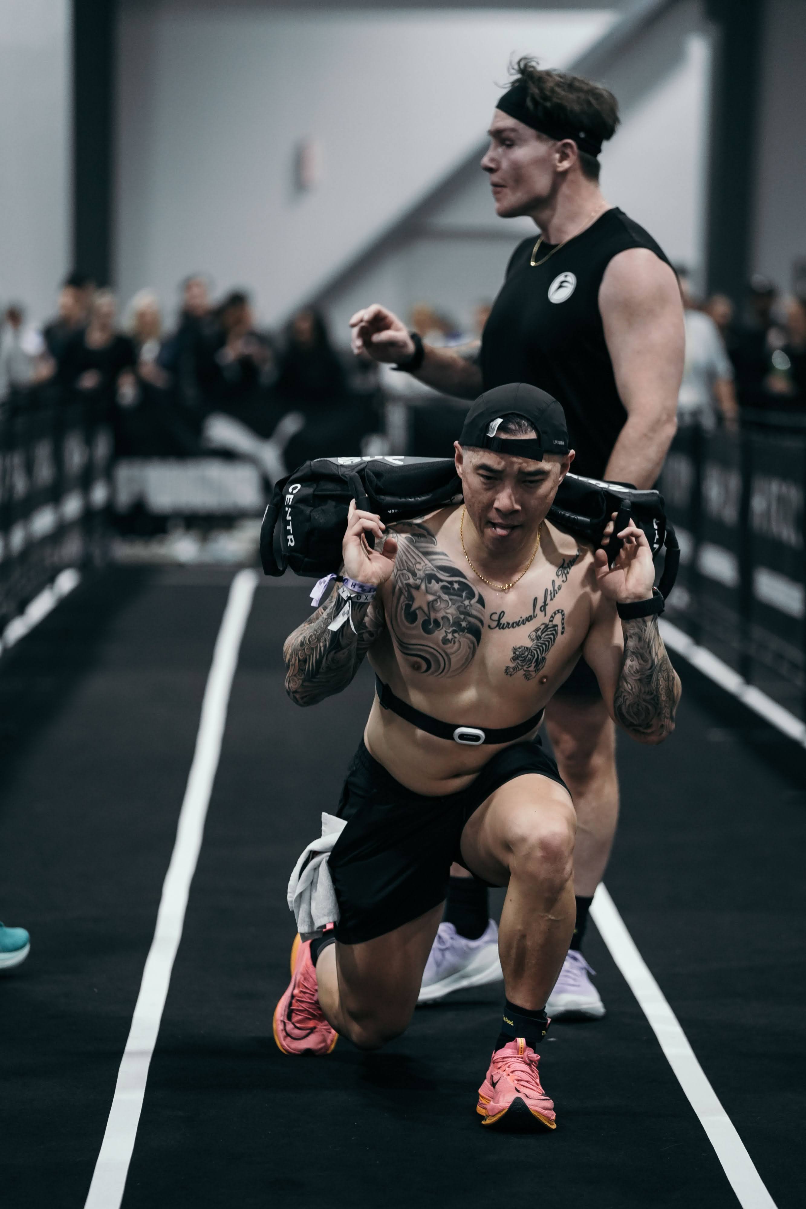 Joe Hsiung carrying a heavy sandbag across his shoulders while performing lunges at the Hyrox Ottawa 2026 Open Doubles competition. He is mid-lunge on the competition floor inside the EY Centre arena with spectators and Hyrox branding visible in the background.