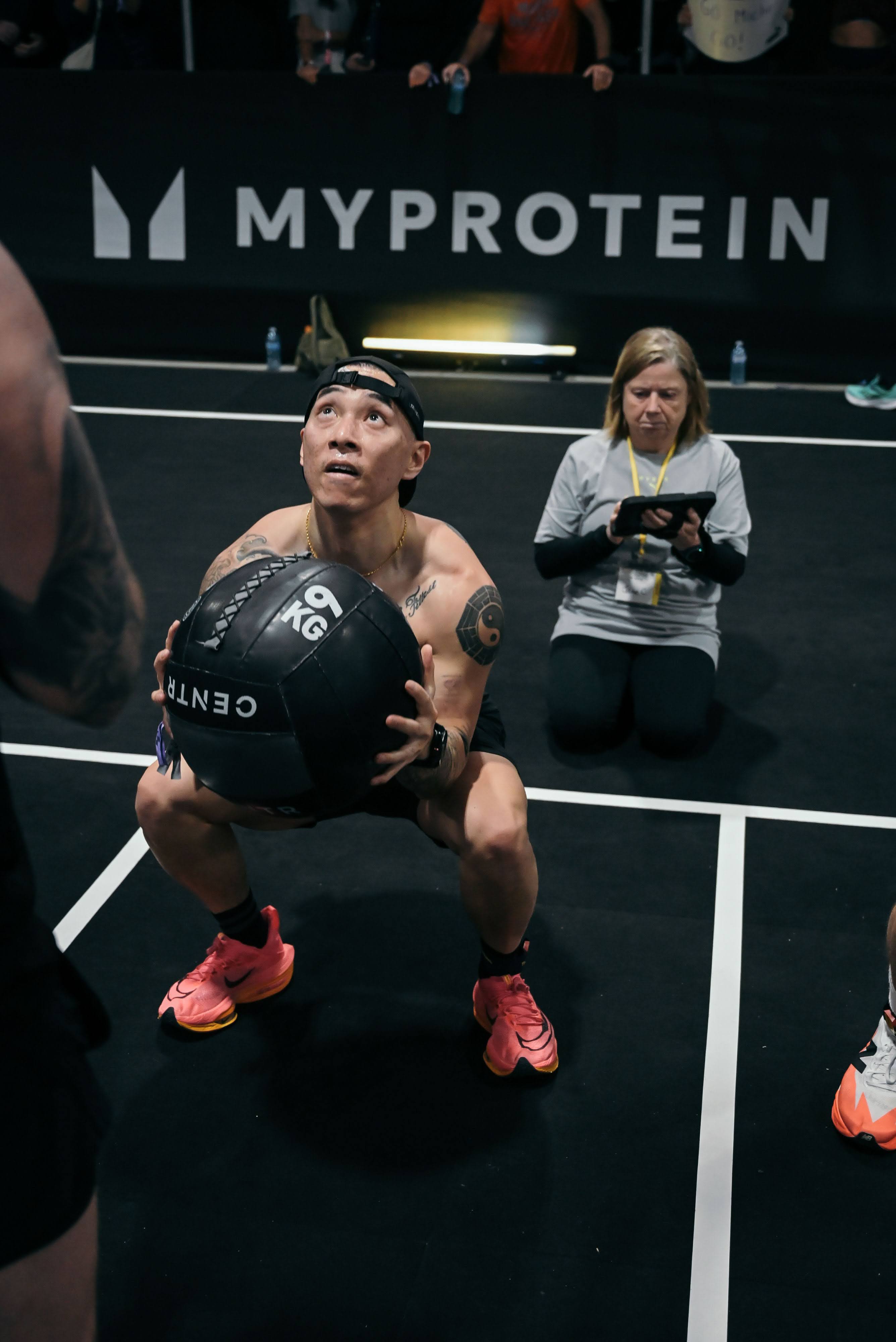 Joe Hsiung performing wall balls at the Hyrox Ottawa 2026 Open Doubles competition. He is in a deep squat position launching a medicine ball upward toward the target on the wall, with a digital rep counter screen visible beside him inside the EY Centre arena.