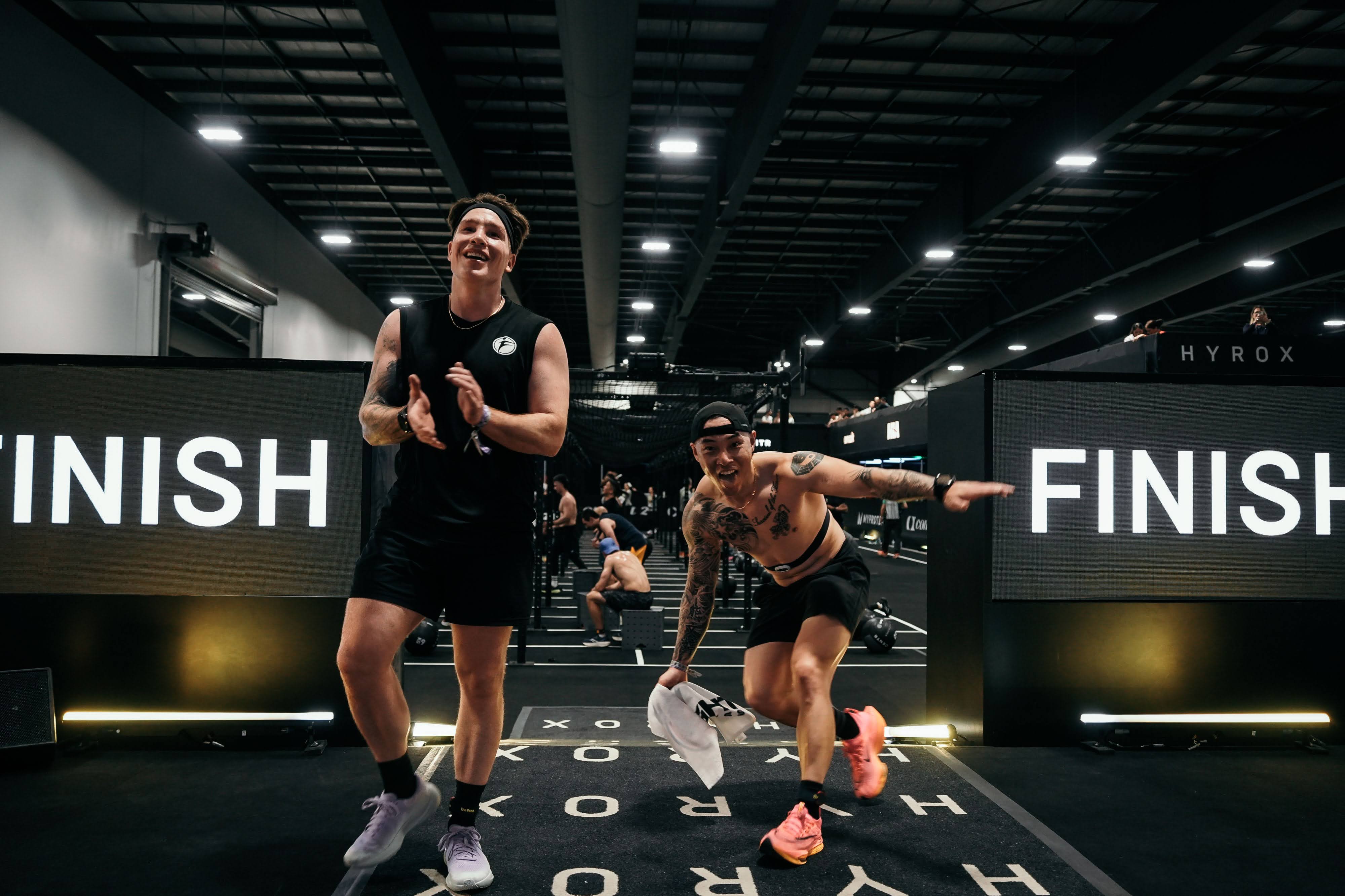 Joe Hsiung and Sean Howard posing together after completing Hyrox Ottawa 2026 Open Doubles. Both are sweaty and exhausted but smiling, holding their finisher patches in front of the Hyrox finish line backdrop inside the EY Centre arena.