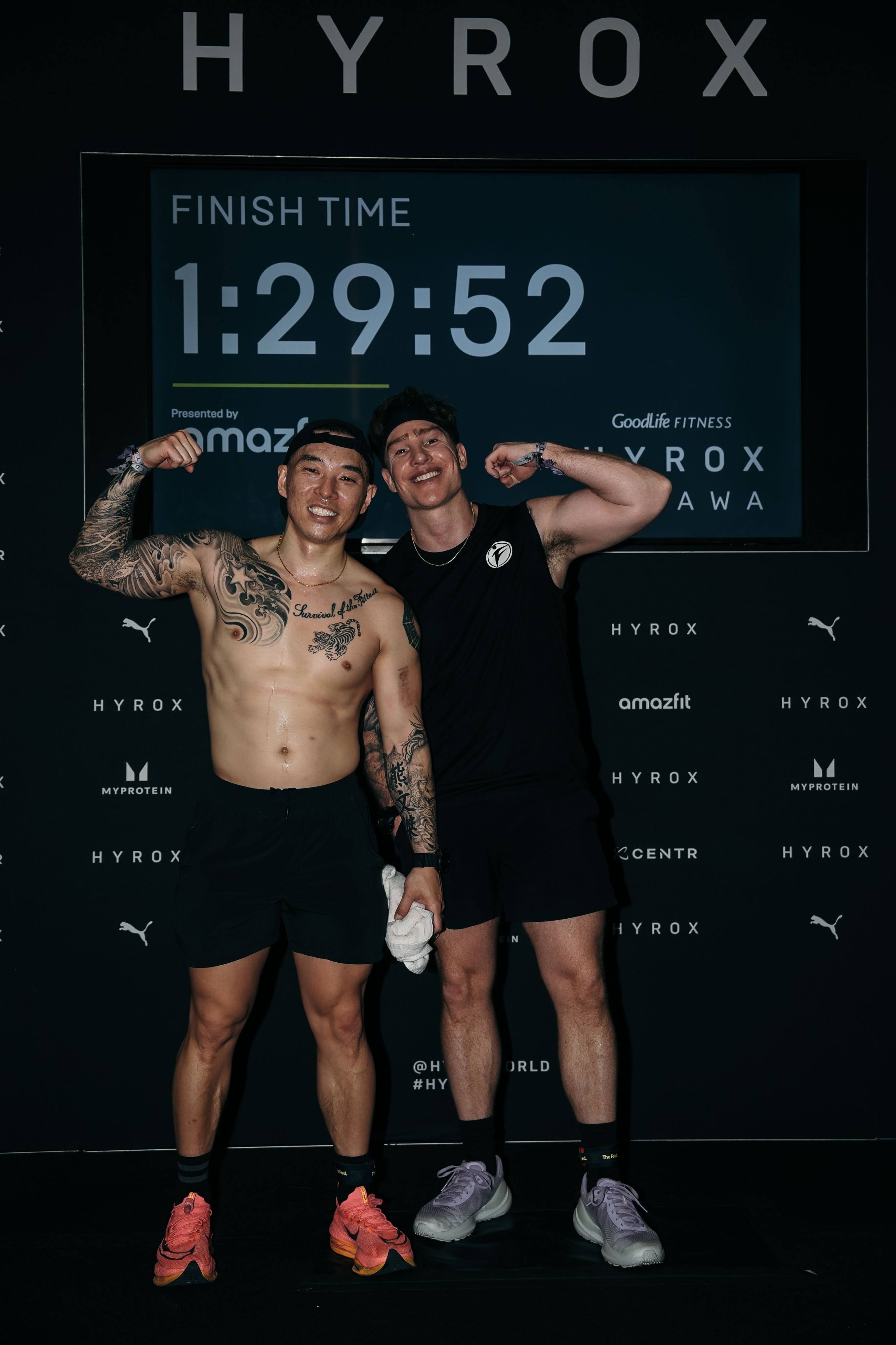 Joe Hsiung and Sean Howard flexing their biceps in front of the Hyrox results screen displaying their finish time of 1:29:52 at the Hyrox Ottawa 2026 Open Doubles competition inside the EY Centre arena.