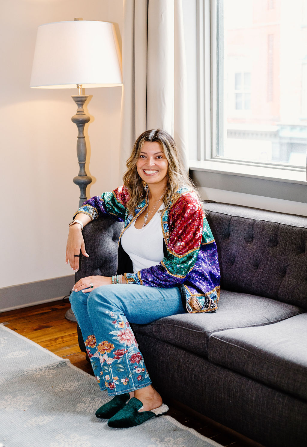 Gigi Bier wearing jeans, white t-shirt and a sequin jacket seating on a sofa smiling.