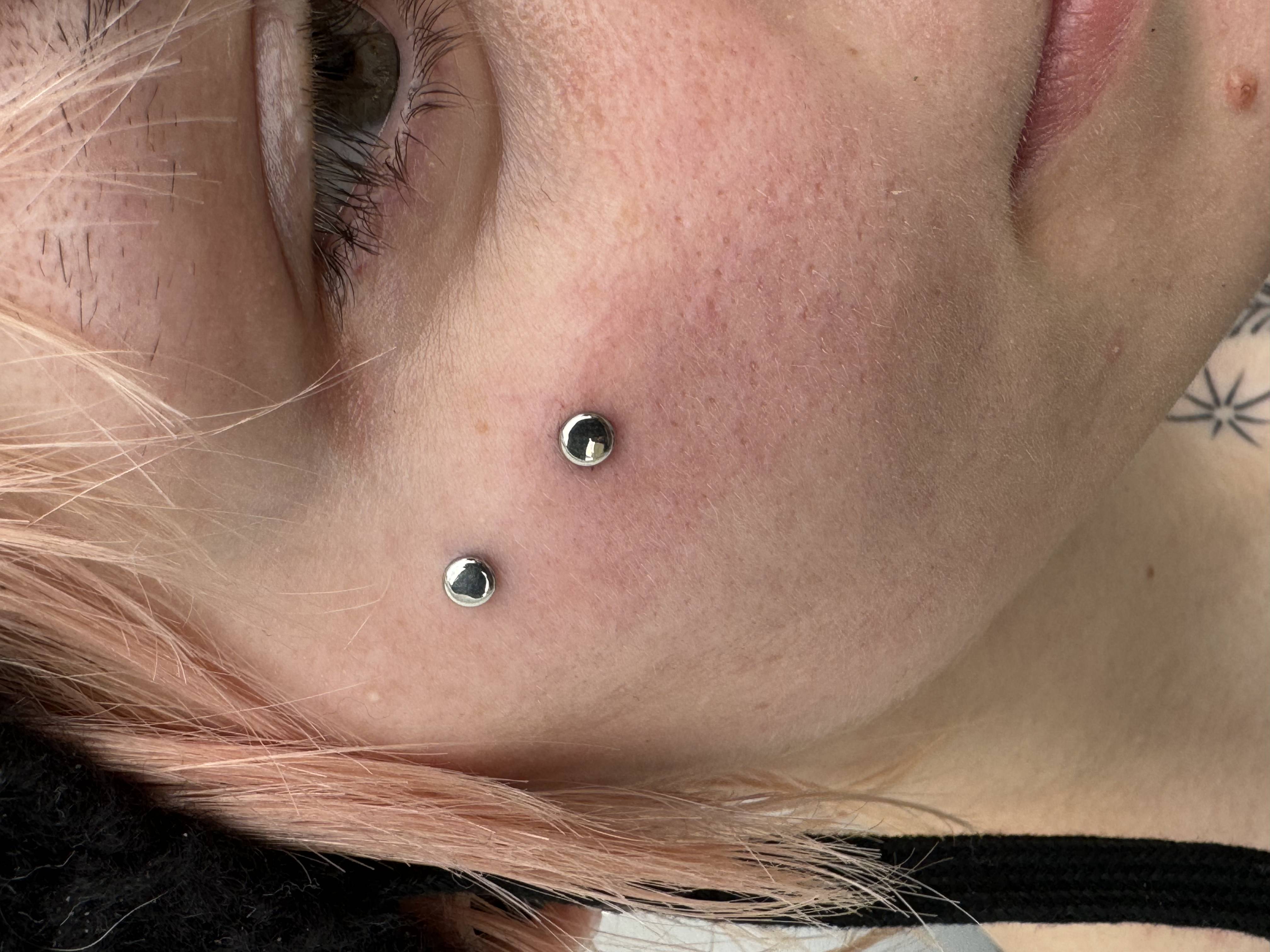 Close-up of a person's face featuring two metallic surface piercings on the cheek, showcasing contemporary body modification artistry by a skilled piercer.