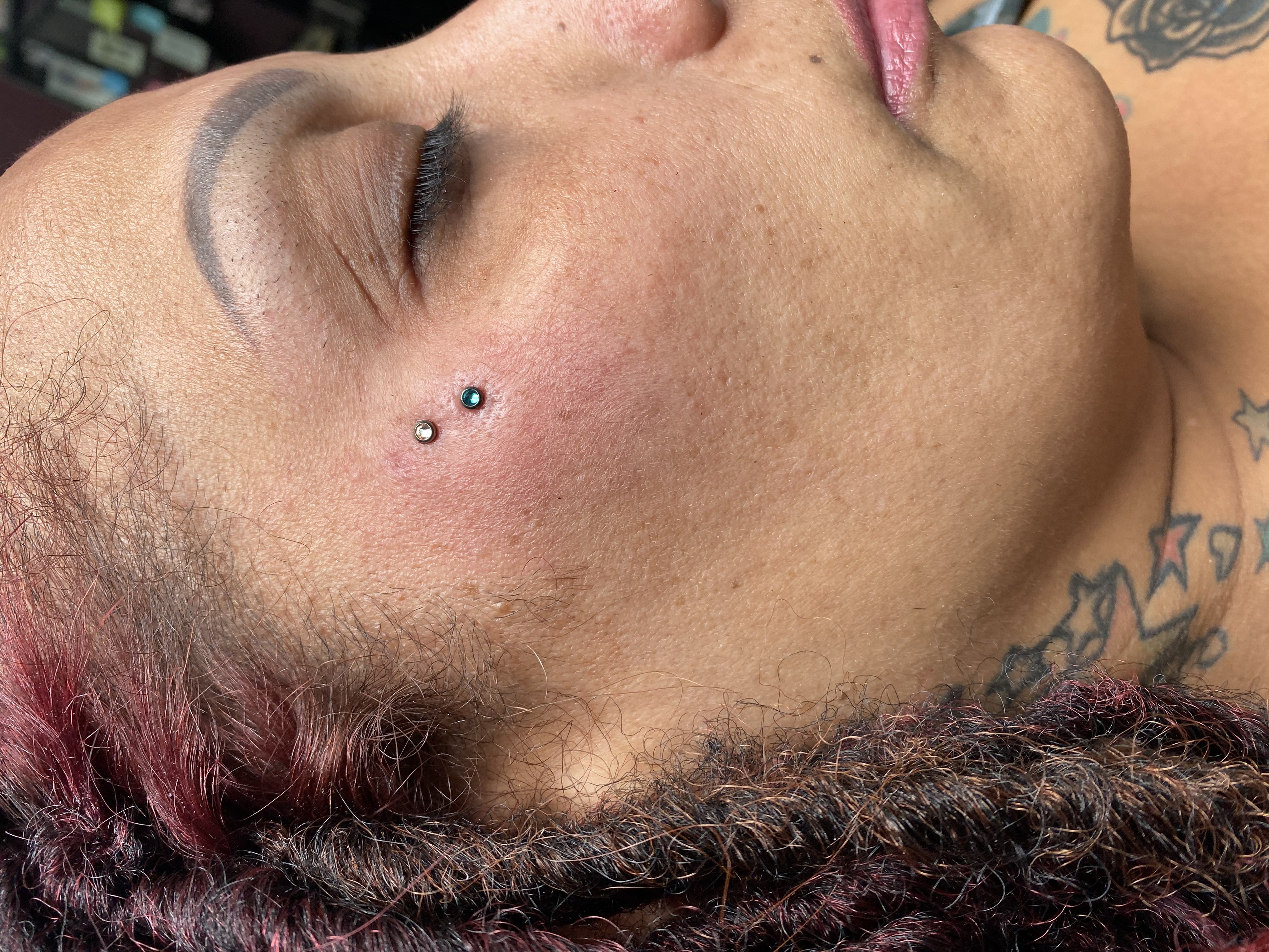 Close-up of a woman with facial piercings, featuring two decorative jewelry pieces on her cheek, showcasing body modification artistry by Mariano at GameFace Tattoos.