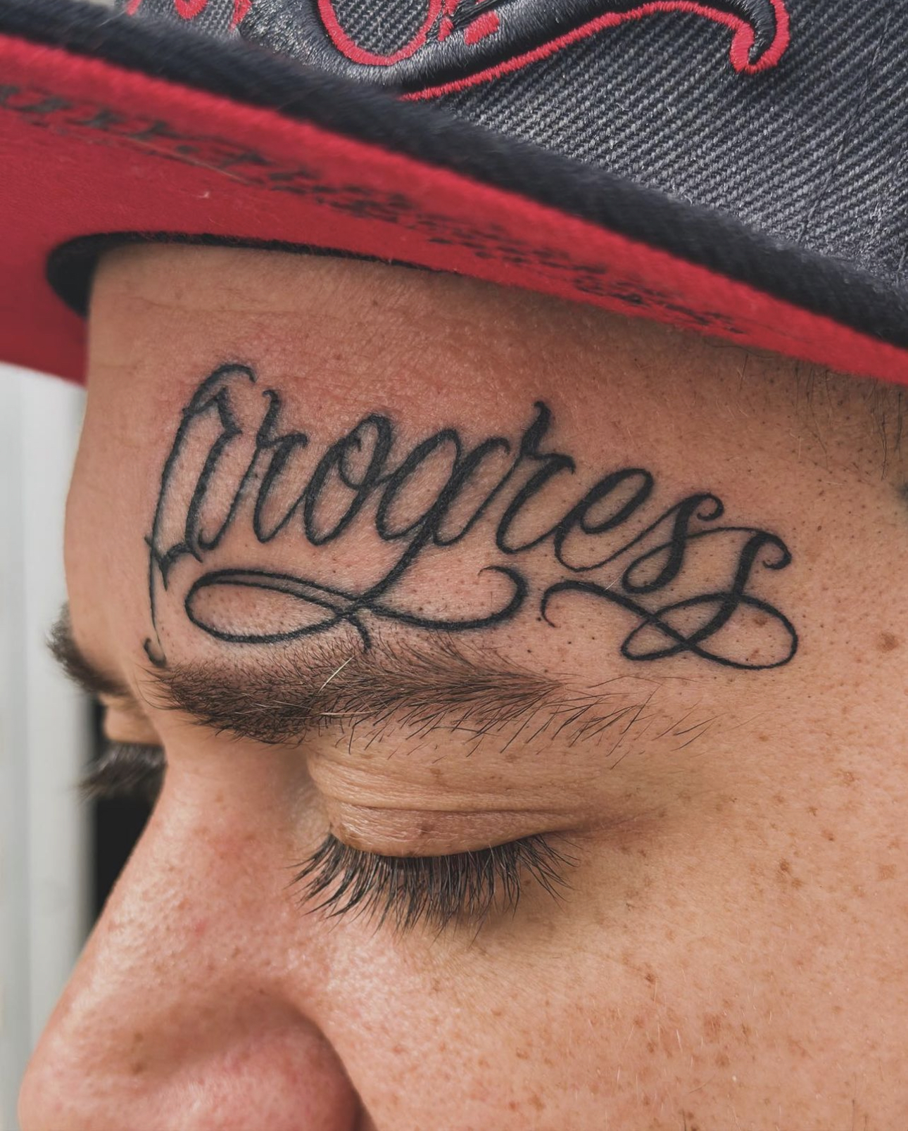 Tattoo of the word "Progress" in cursive on a person's forehead, showcasing detailed ink work and a close-up view of the individual's face, emphasizing the artistry of tattoo culture.