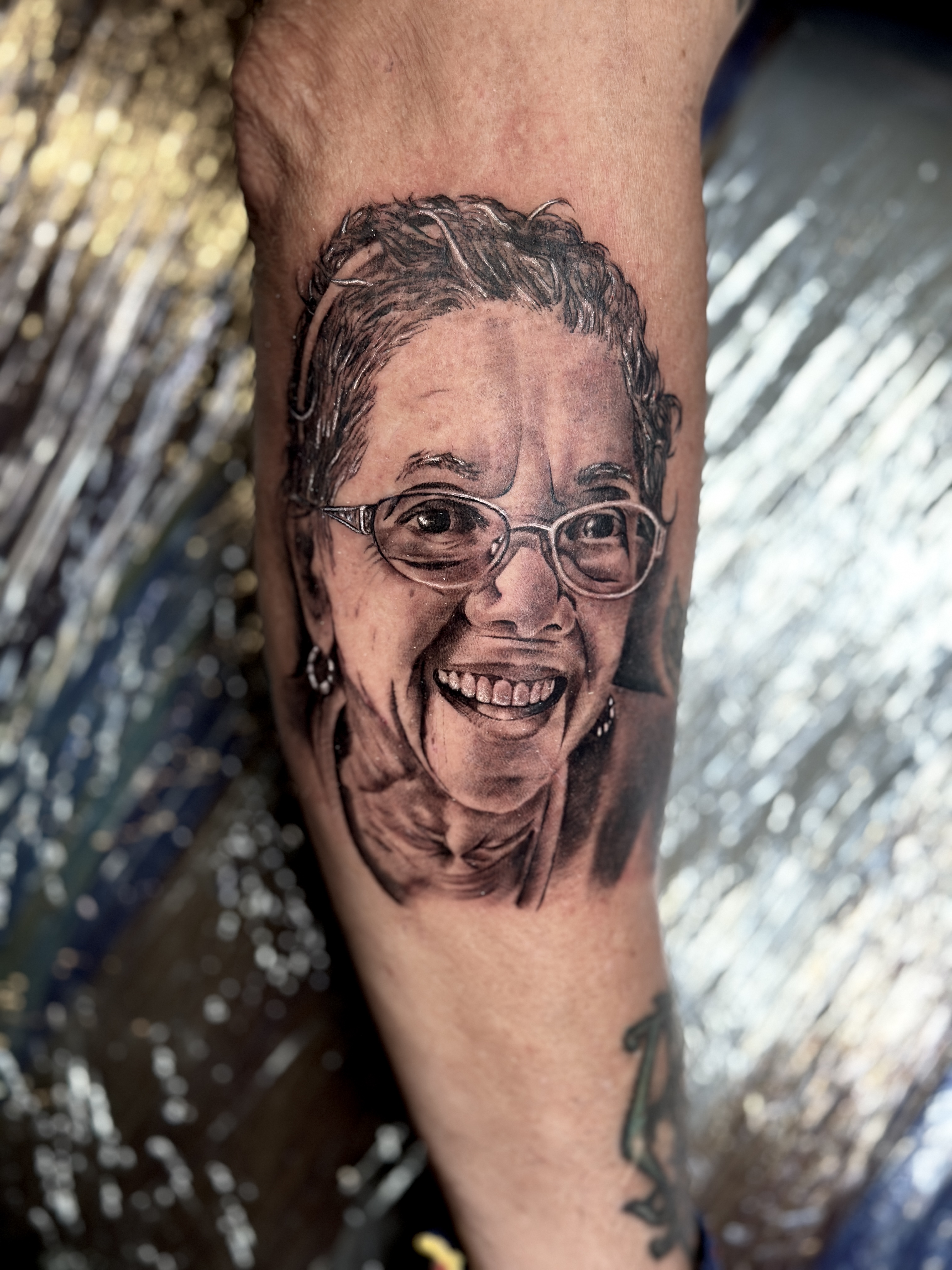 Tattoo of a smiling elderly woman with glasses, showcasing detailed artistry and emotional expression, reflecting personal stories and identity in tattoo design.