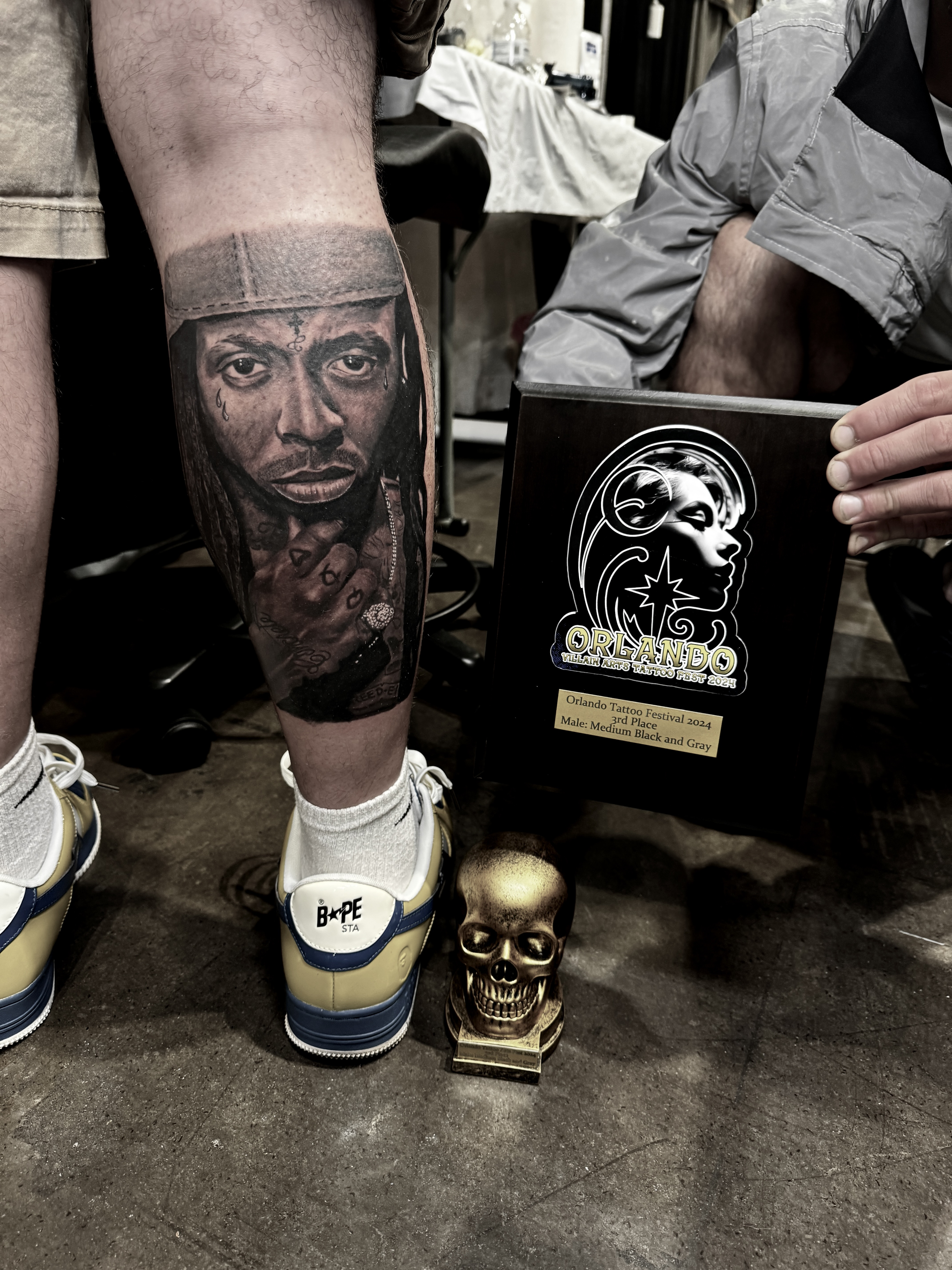 Tattoo of a detailed portrait on a leg, showcasing intricate shading and realism, with an award plaque and a small golden skull in the foreground, reflecting the artistry and recognition in the tattooing community.