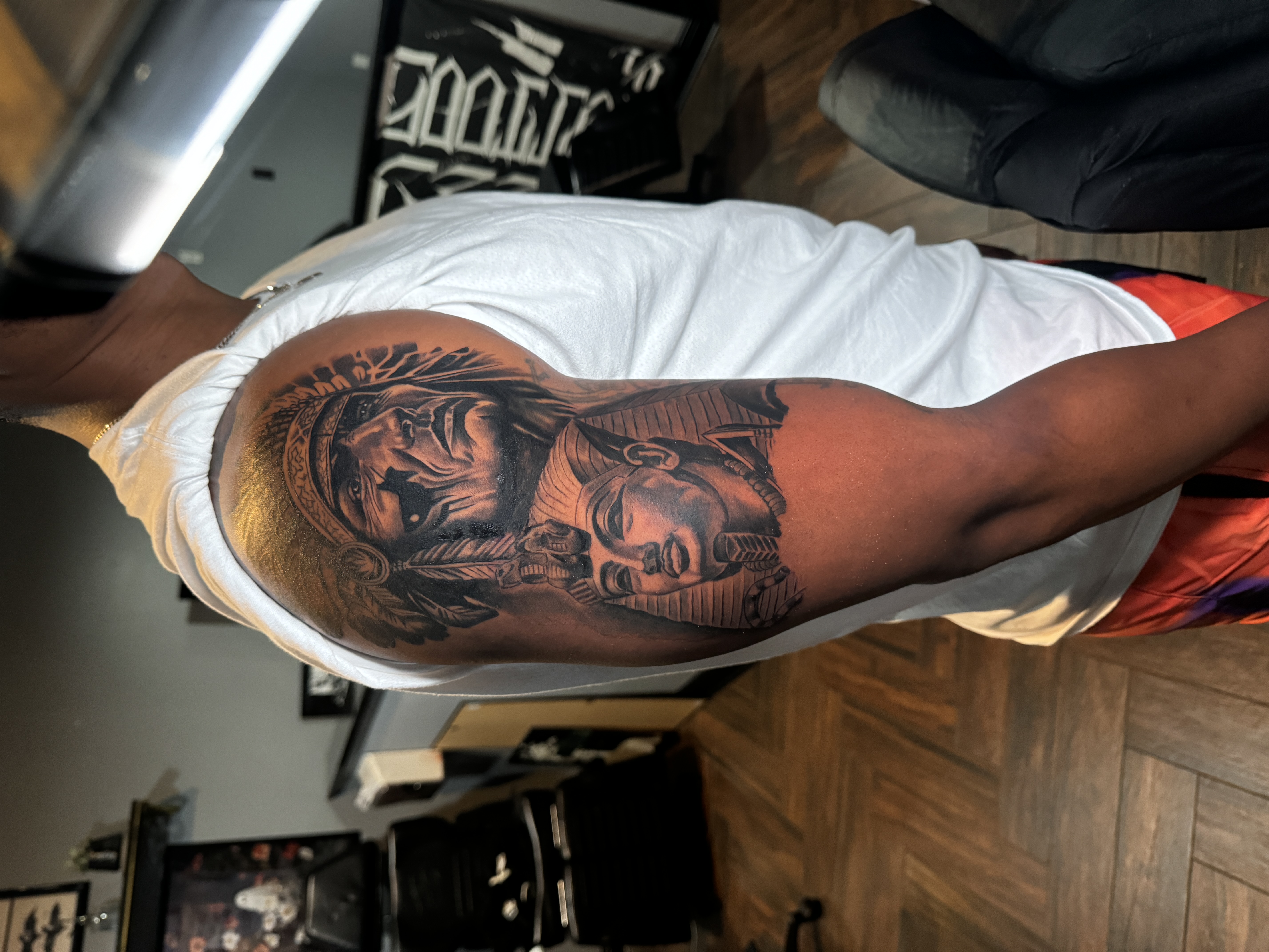 Tattoo of a Native American chief and an Egyptian pharaoh on a man's upper arm, showcasing intricate details and artistry in a tattoo studio setting.