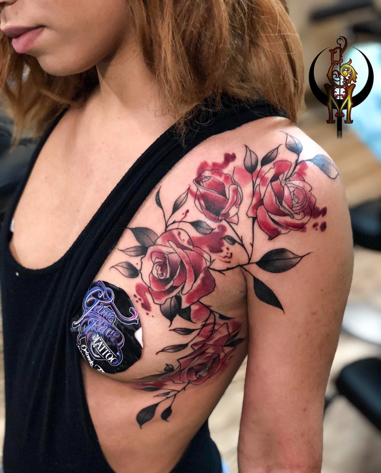 Tattoo of red roses and black leaves on a woman's shoulder, showcasing intricate details and vibrant colors, with a GameFace Tattoo logo visible on the skin.