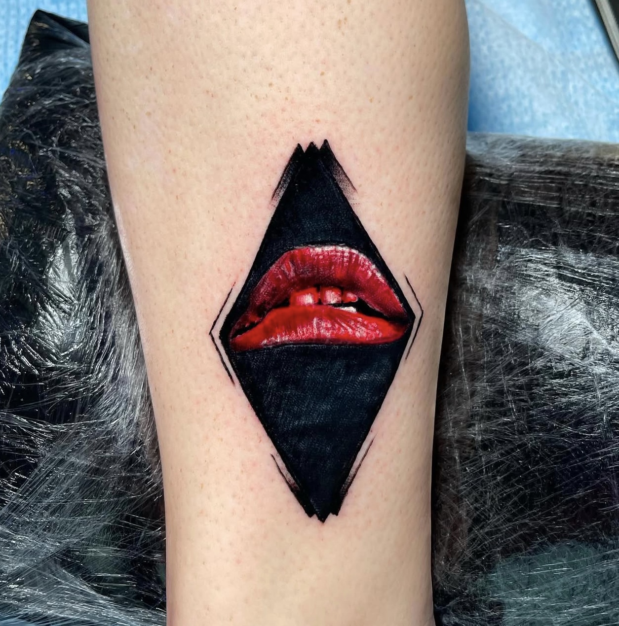 Tattoo of stylized red lips within a black diamond shape on a person's forearm, showcasing Edward Madera's unique artistry at GameFace Tattoo.