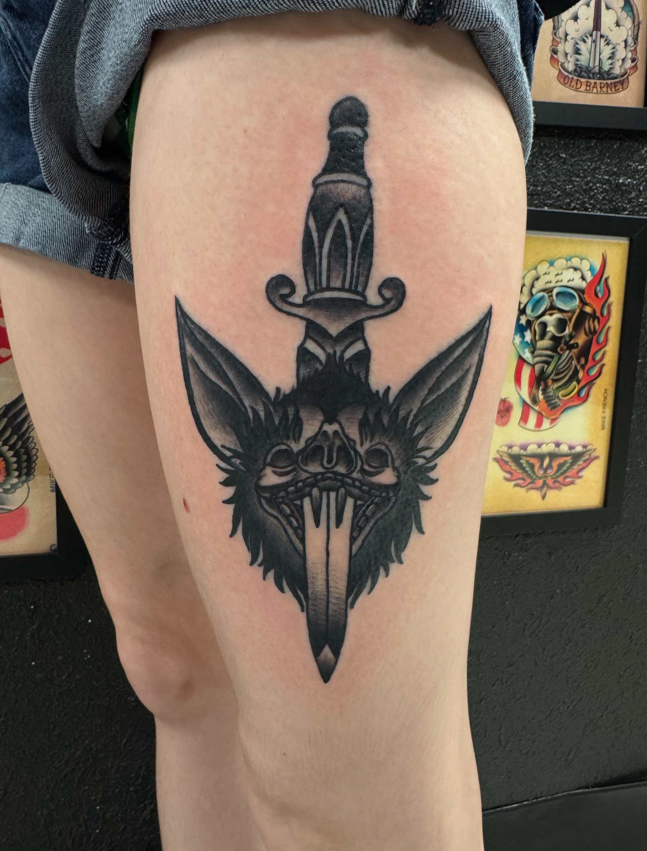 Tattoo of a dagger with a stylized animal face, featuring intricate detailing and bold black ink, displayed on a thigh against a backdrop of tattoo art.