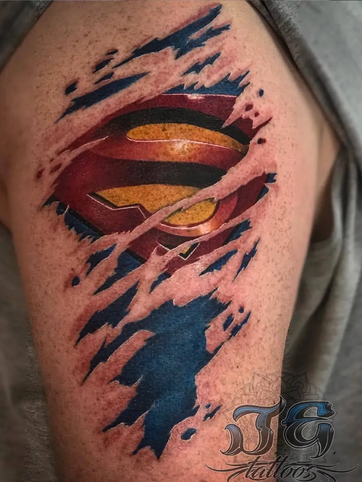 Tattoo design featuring a realistic Superman logo emerging from torn skin, showcasing vibrant colors and intricate shading, highlighting Game Face Tattoo's expertise in comic-inspired artwork.