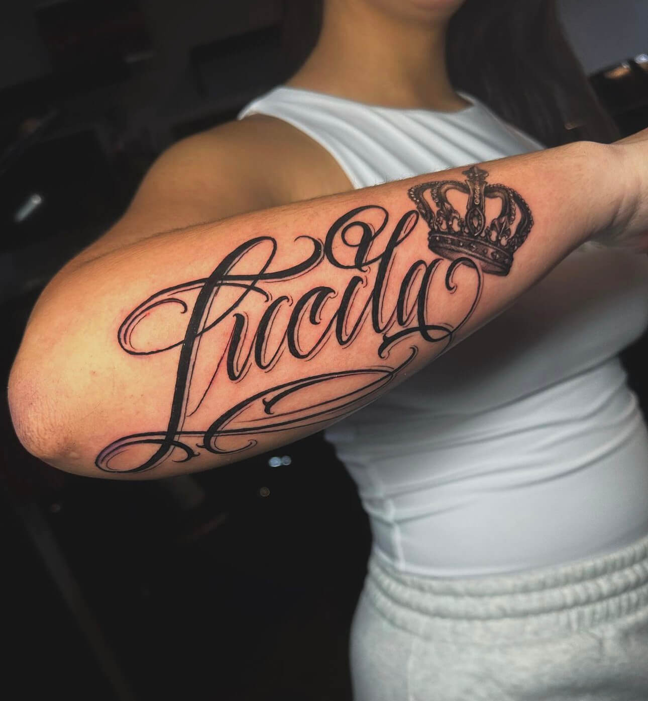 Tattoo of the name "Lucila" in elegant script with a crown above, showcasing black and grey ink on a woman's forearm, reflecting personalized tattoo artistry.