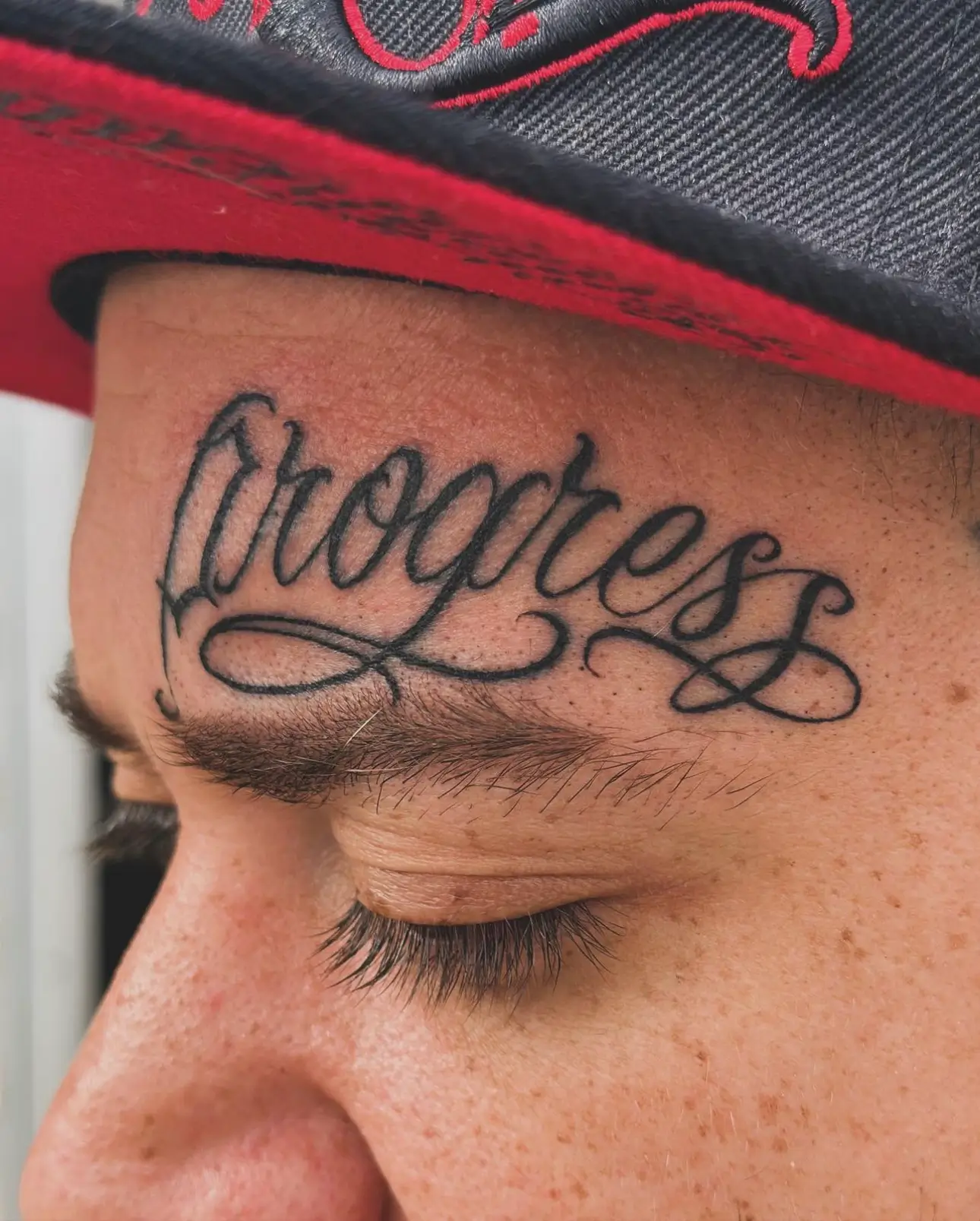 Tattoo of the word "Progress" in elegant script on a person's forehead, showcasing detailed artistry and shading, relevant to GameFace Tattoo's portfolio of unique designs.