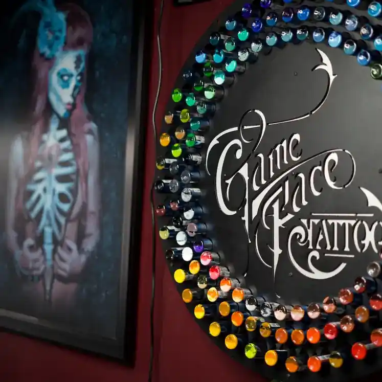 GameFace Tattoos logo displayed on a circular palette of vibrant tattoo ink bottles, with a colorful mural of a skeleton-themed woman in the background, highlighting the studio's artistic flair and creativity.