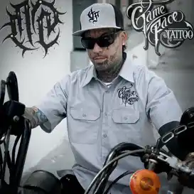 Tattoo artist wearing a gray shirt with GameFace Tattoos logo, sunglasses, and a cap, seated on a motorcycle, showcasing the studio's edgy style and expertise in body art.
