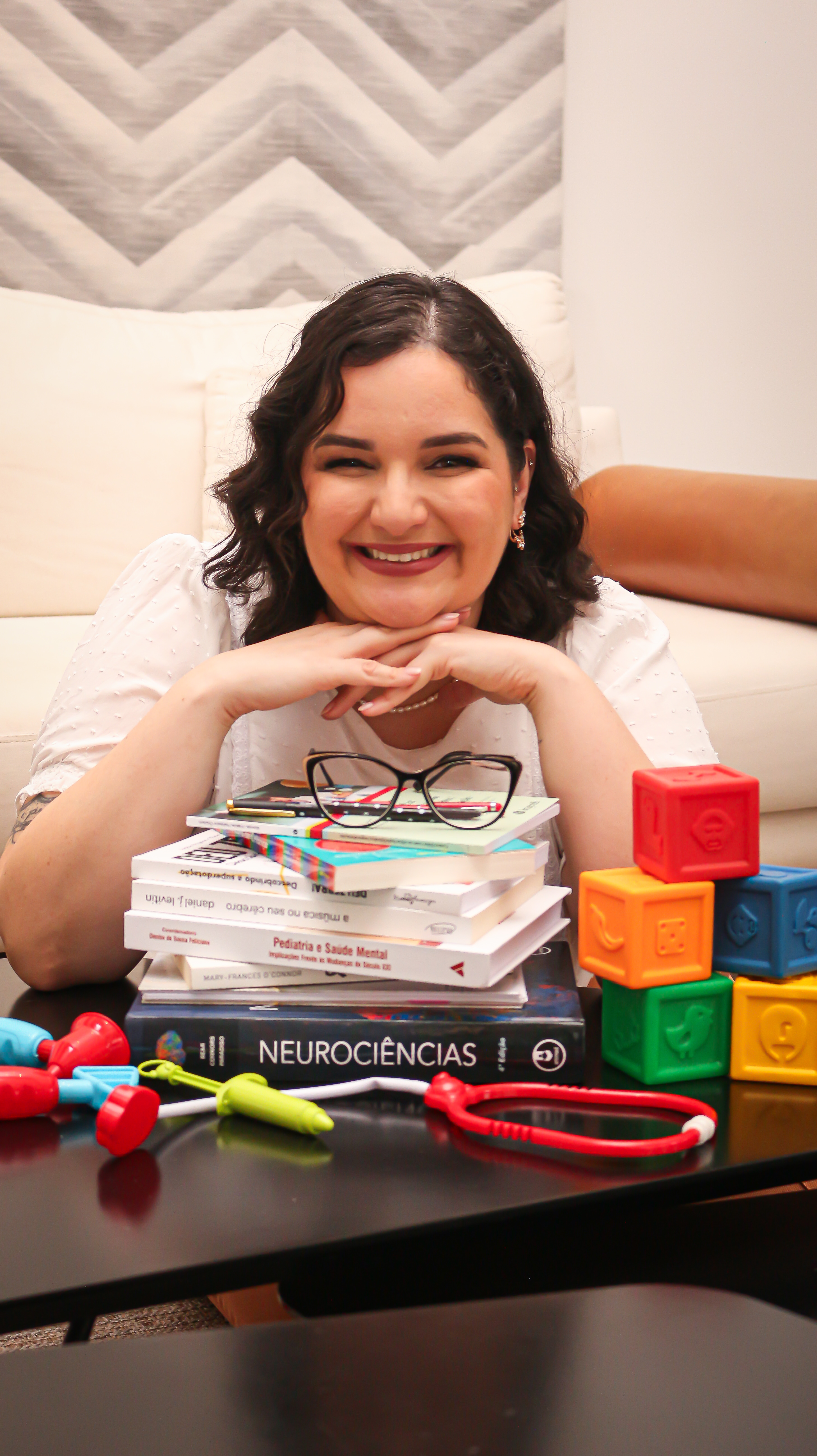 Dra. Romana sorrindo, apoiada em livros, com brinquedos infantis ao lado