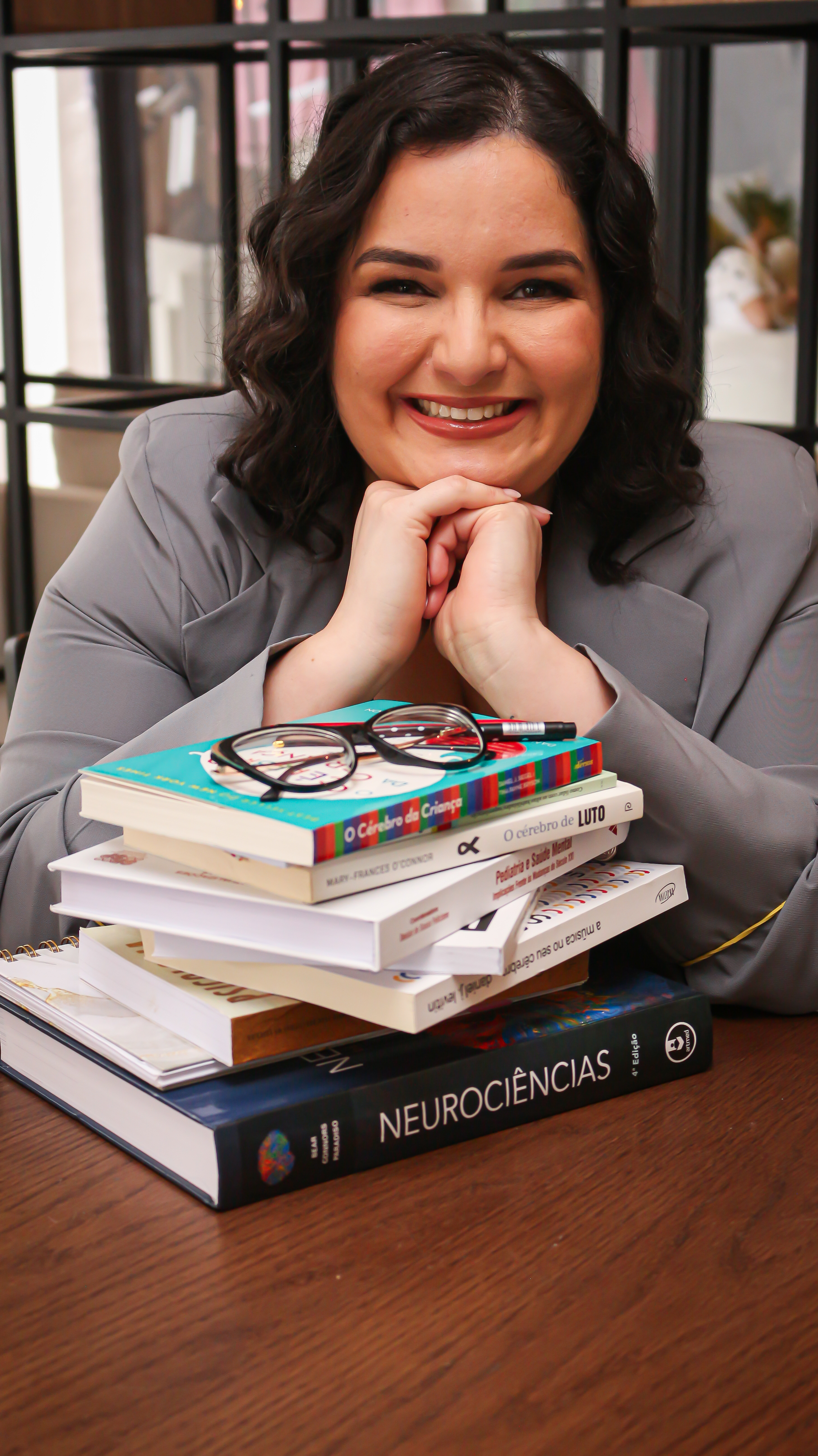 Dra. Romana Medeiros sorrindo atrás de uma pilha de livros sobre infância e neurociências