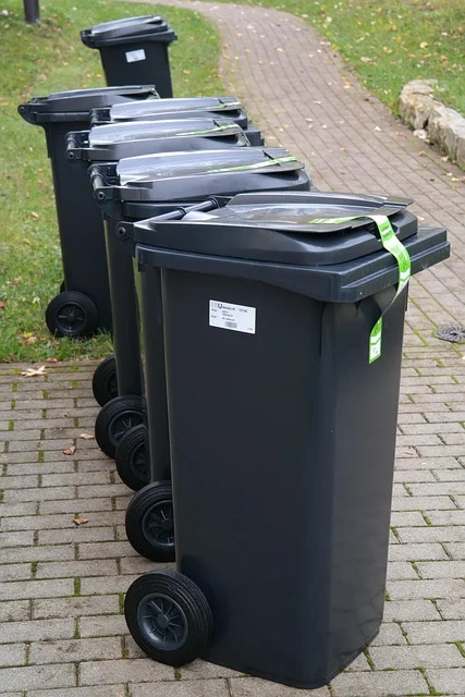 A line of five dark gray recycling bins with lids slightly open and green tags, on a brick pathway.