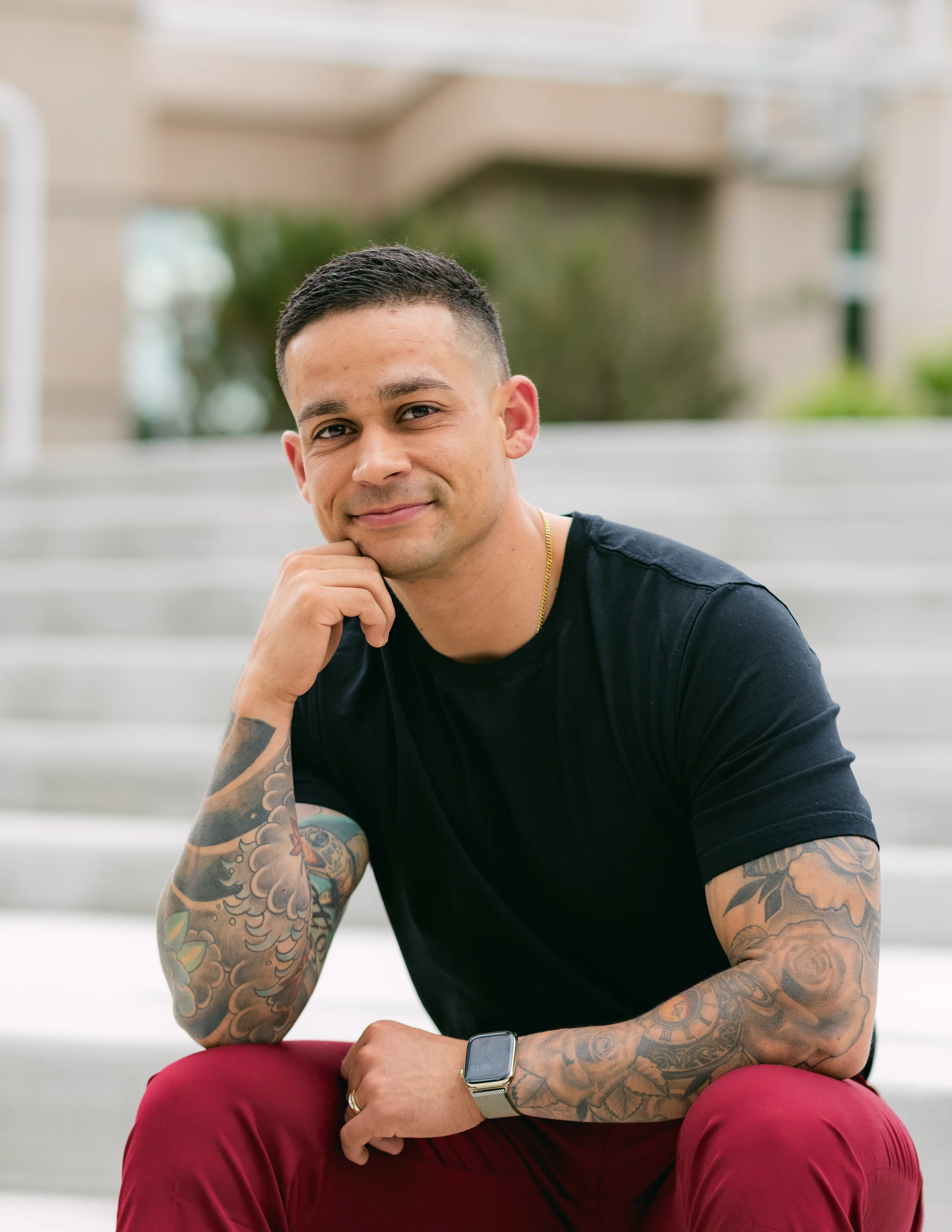 A smiling man with short hair and tattoos on his arms, dressed in a black t-shirt and red pants, sits on outdoor steps.