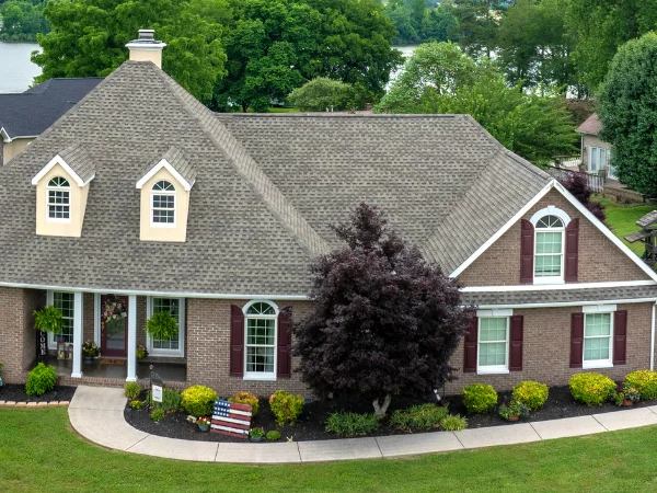 An aerial view of a large brick house with a brown shingle roof, dormer windows, and dark red shutters, overlooking a river.