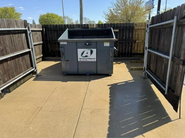 A gray Advanced Disposal dumpster sits in a fenced enclosure with wooden panels and metal posts on a concrete pad.