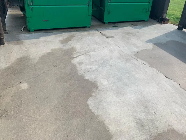 Patches of wet concrete with cracks on a ground under two large green dumpsters, with sun visible on the right.