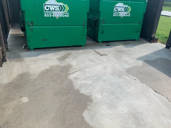 Two large green dumpsters from CWS Capital Waste Services standing on a wet concrete pad with various cracks.