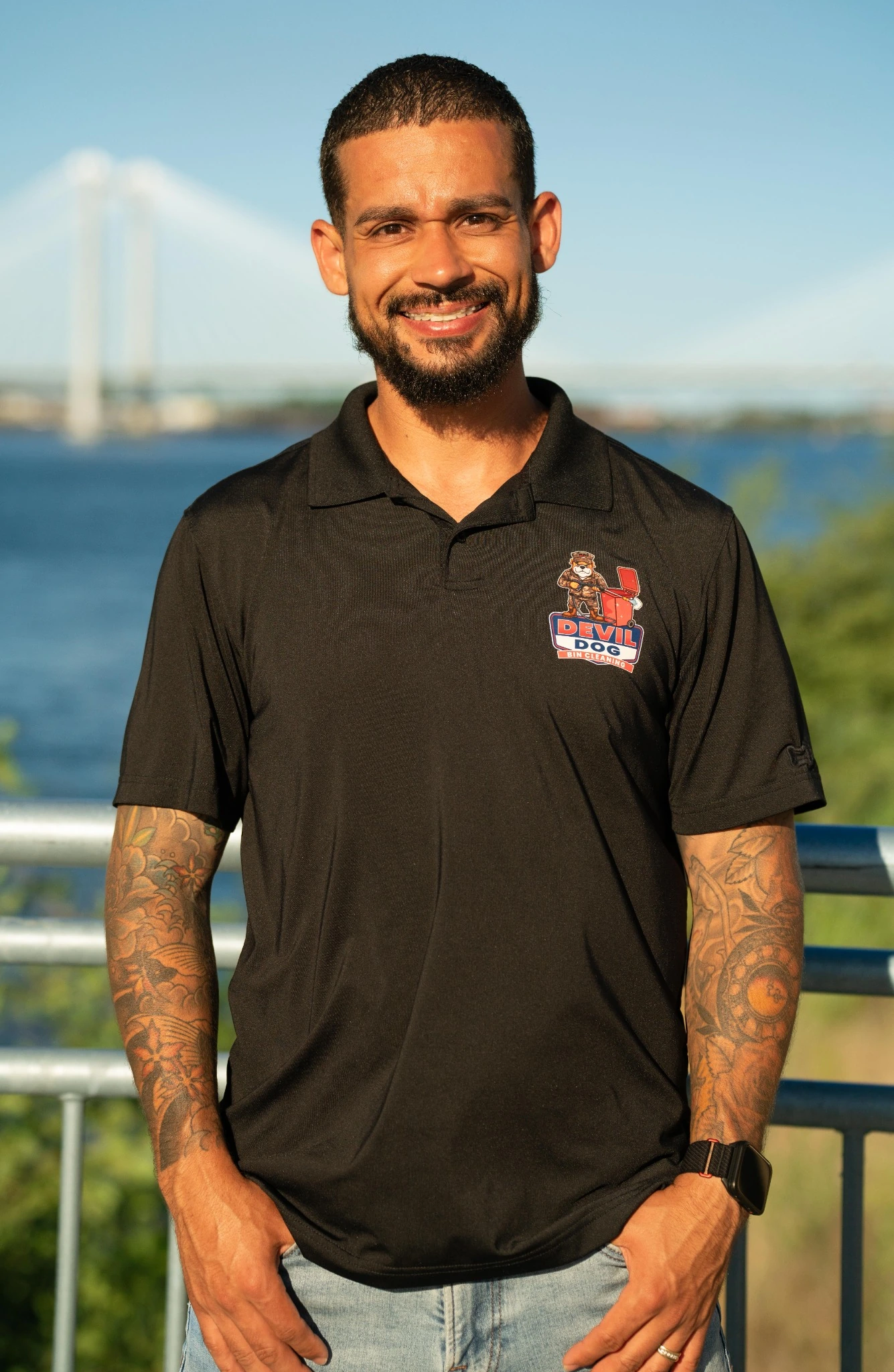 A smiling man in a black polo shirt with a Devil Dog Bin Cleaning logo, blue jeans, and tattoos stands outdoors.