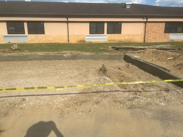 Partially constructed or unrenovated railroad industrial site with a long, narrow concrete trench running through the center. The trench is bordered by raised concrete edges, and the surrounding area appears to be a warehouse or a similar large indoor space. Scaffolding, construction equipment, and safety barriers are visible in the background, indicating ongoing construction or maintenance work.