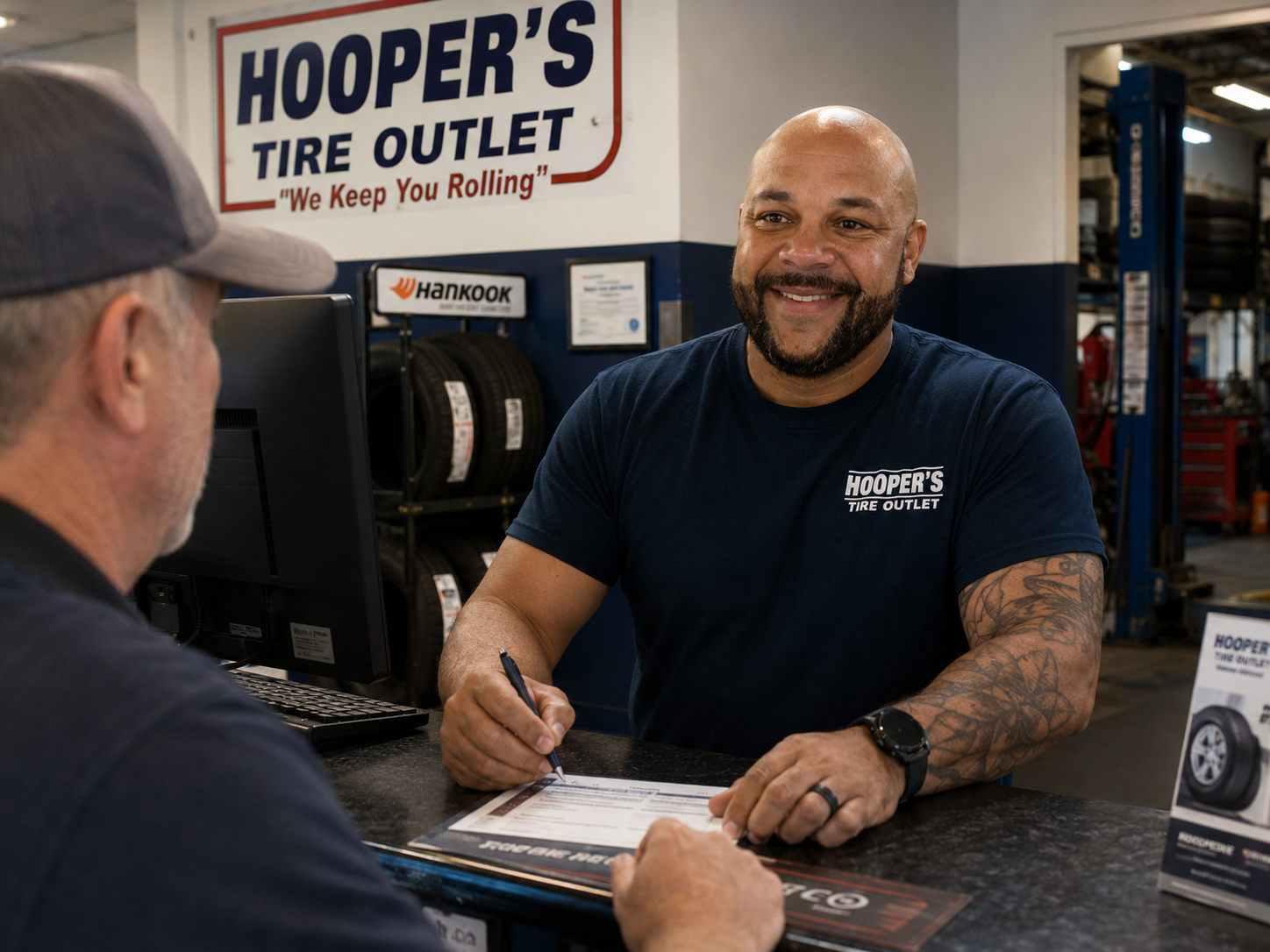 A Hooper's Tire Outlet team member smiling at the counter while writing up a work order for a customer, with the service bays and tire racks visible behind him.