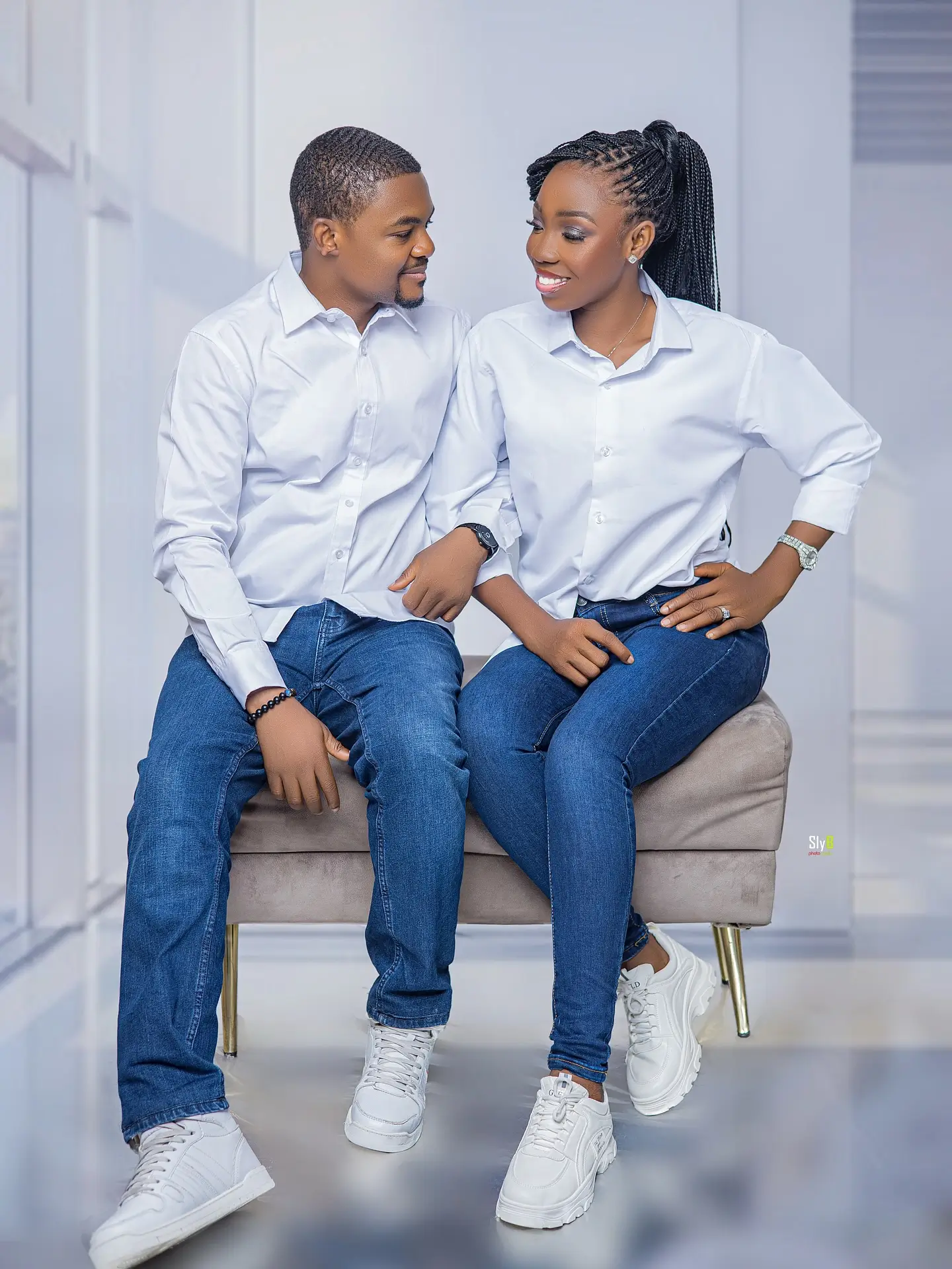 Couple in white shirts and jeans seated on a bench looking at each other and smiling.