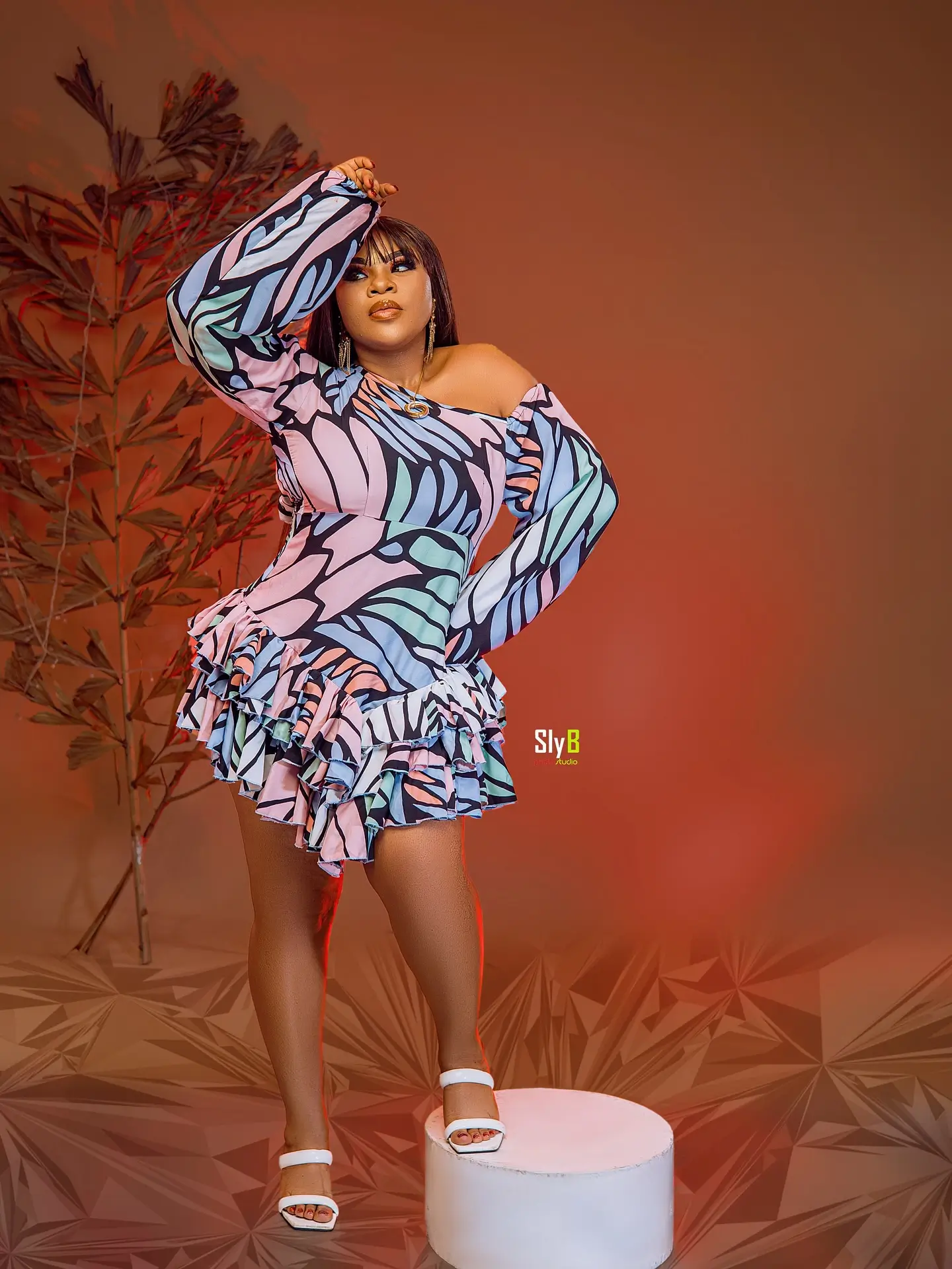 Woman in a colorful patterned dress posing with one arm raised against a red backdrop.