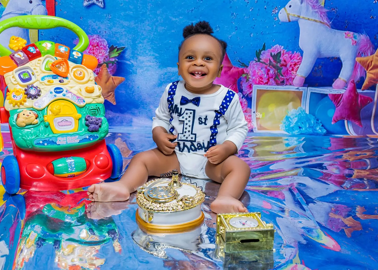 Baby in a first birthday onesie with a bow tie sitting among toys and a unicorn backdrop.