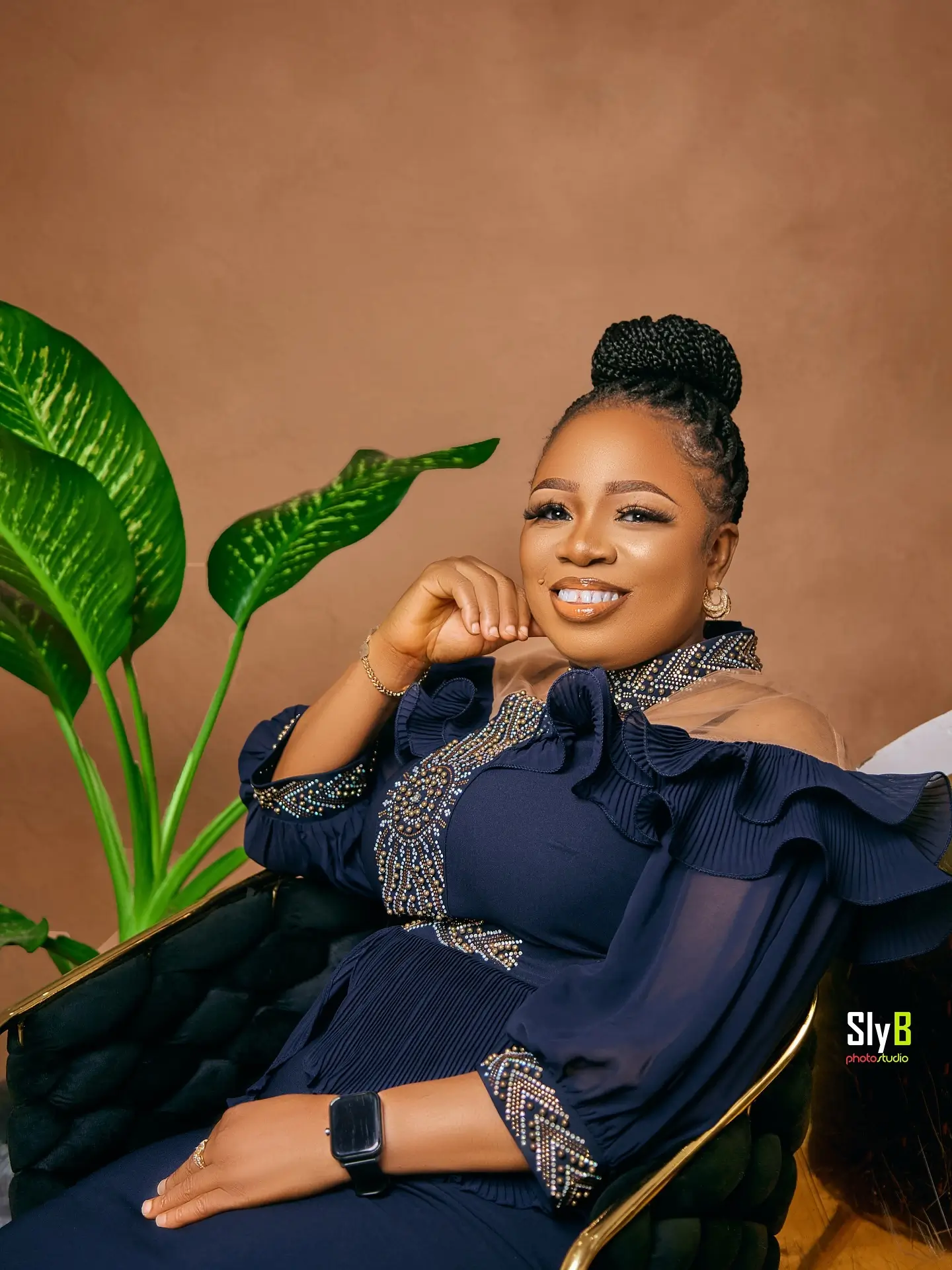 Woman in a navy ruffled outfit seated in a chair and smiling beside a plant.