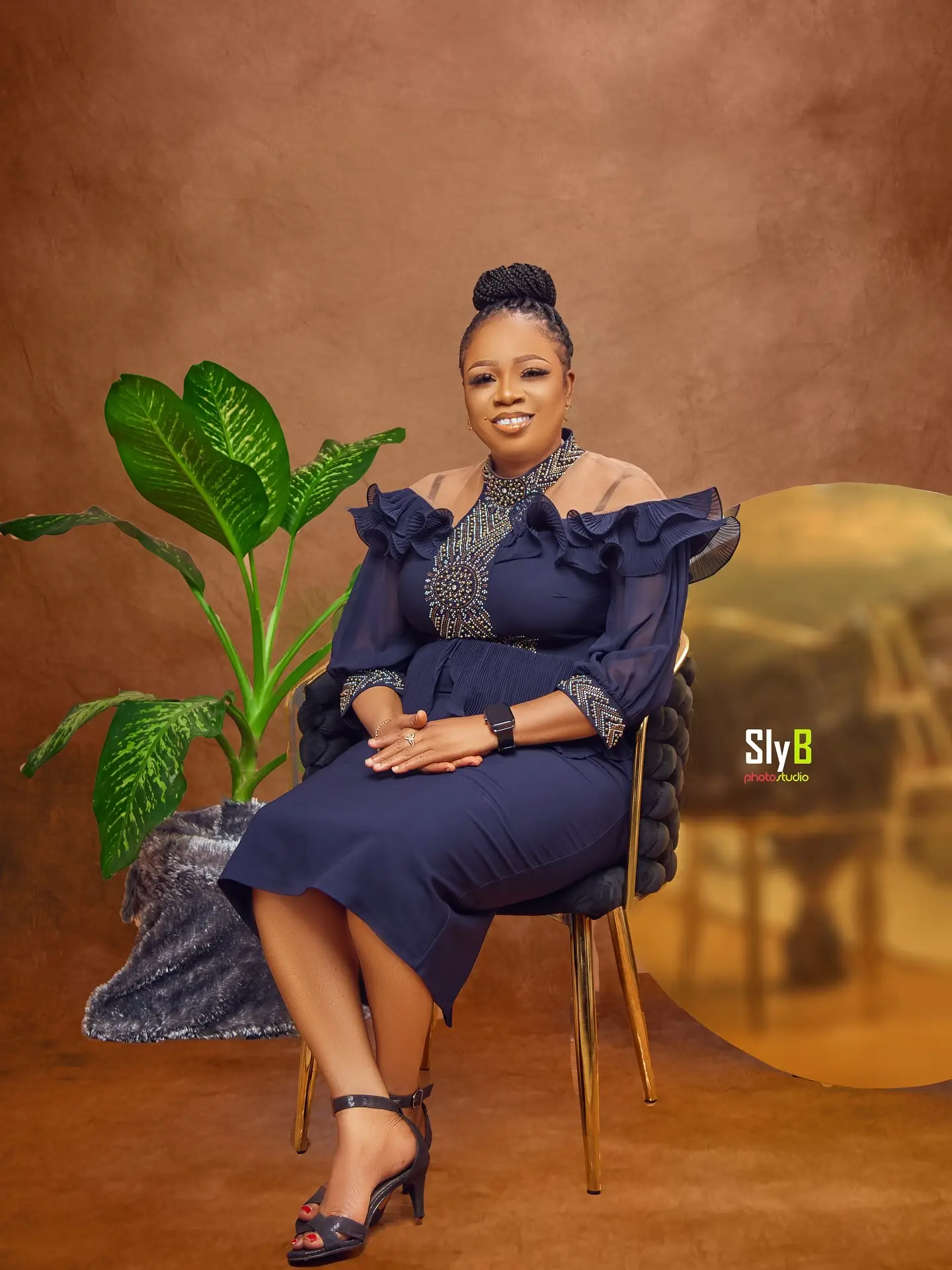 Woman in a navy ruffled outfit seated in a chair beside a leafy plant.
