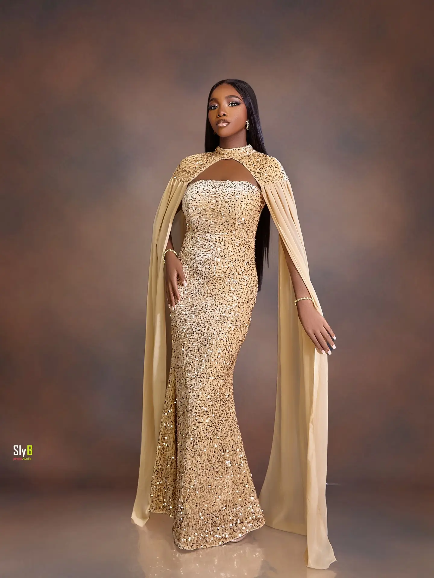 Woman in a gold sequin gown with cape sleeves standing against a brown backdrop.