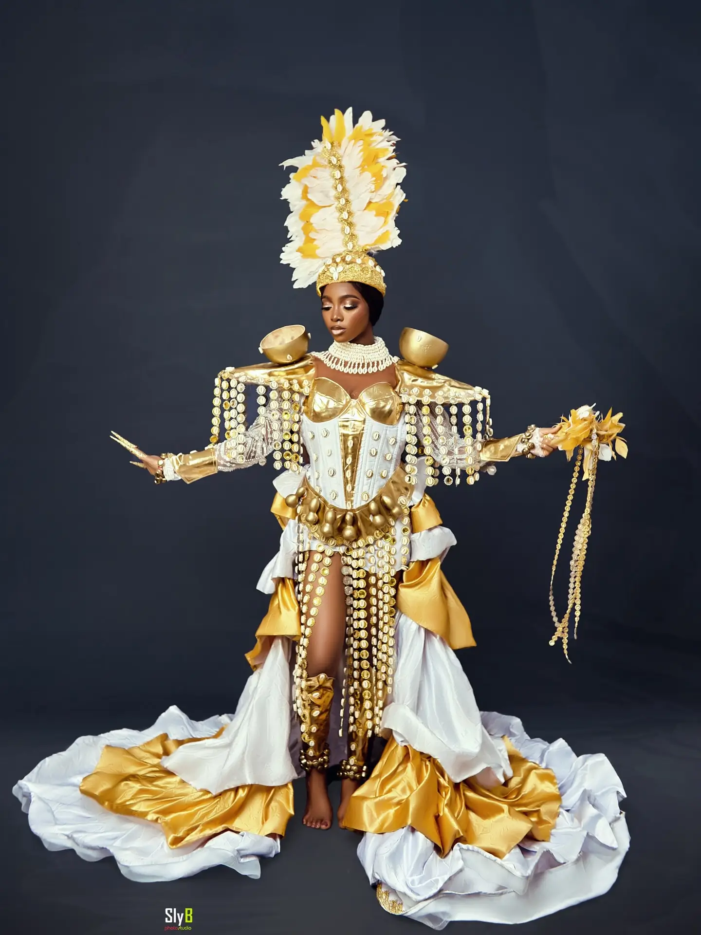 Woman in an elaborate gold and white ceremonial costume with a feather headdress and cowrie shell beads.