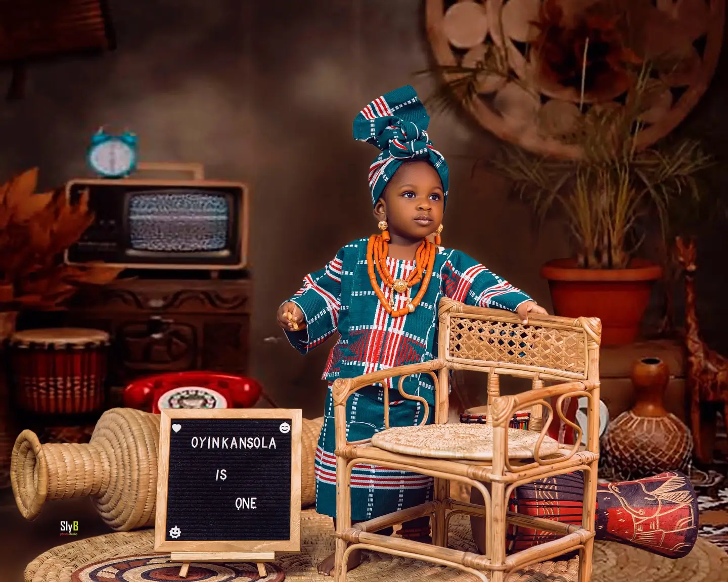 Toddler girl in traditional attire standing beside a sign reading Oyinkansola is one.