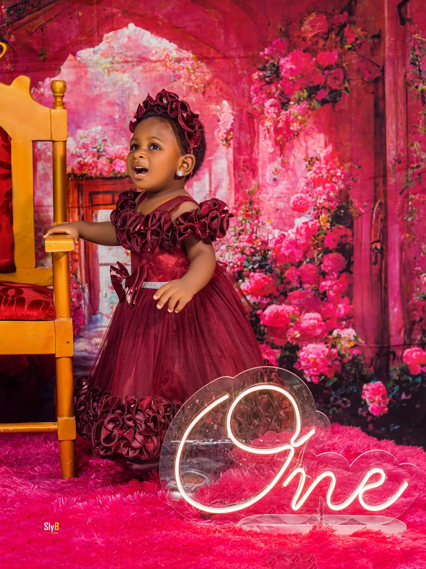 Toddler girl in a maroon dress posing near a floral backdrop for her first birthday.